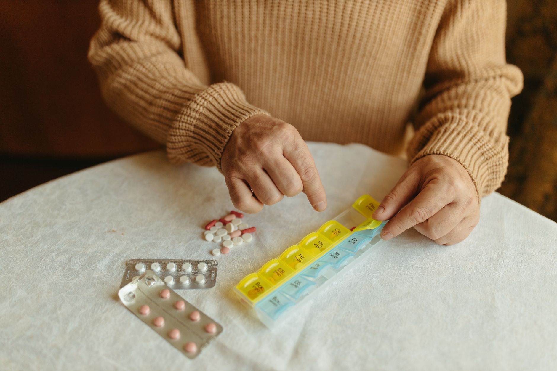 Hands organizing various pills into a weekly pill organizer on a table.