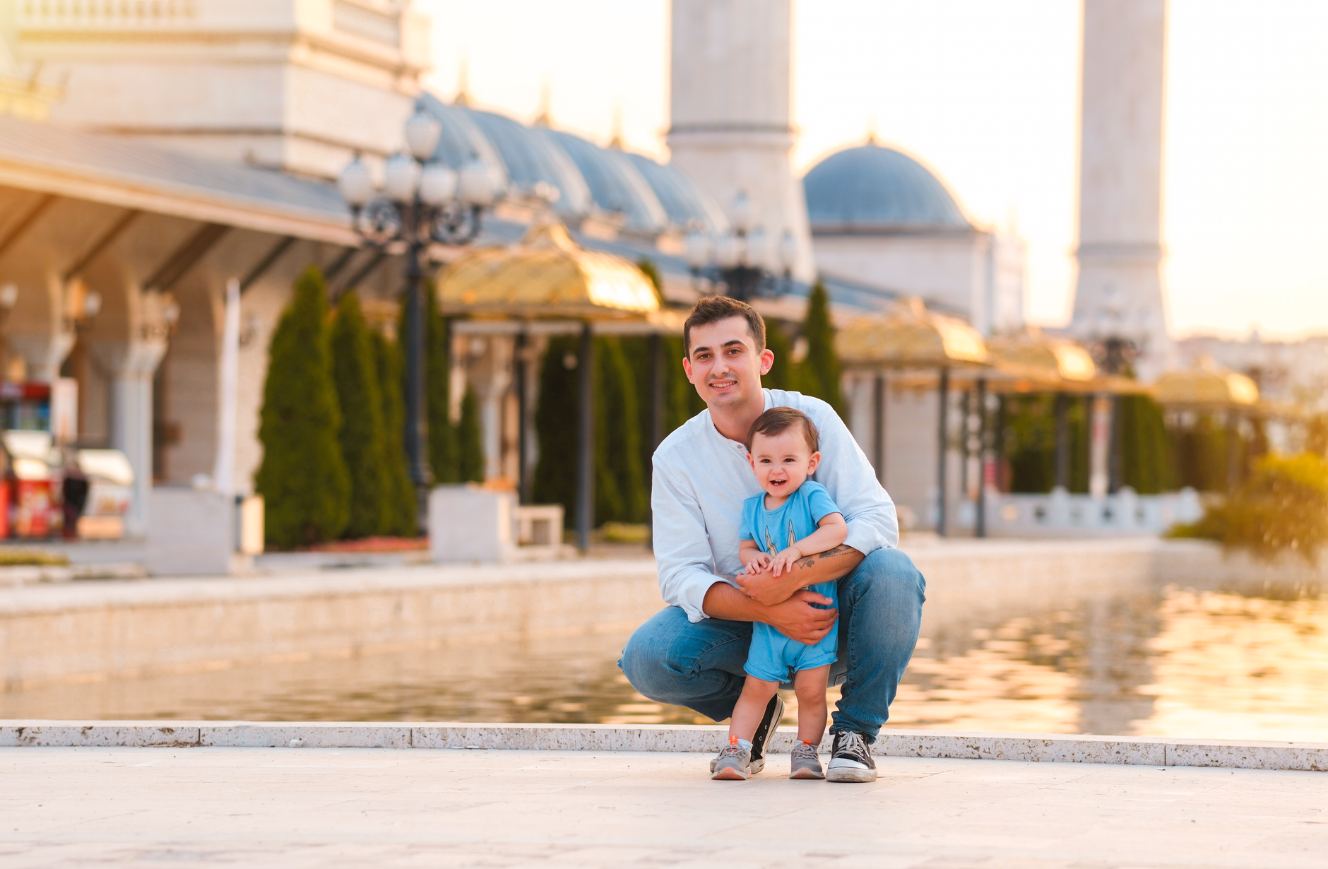 A man kneels beside a happy toddler near a reflective water surface in Türkiye during sunset.