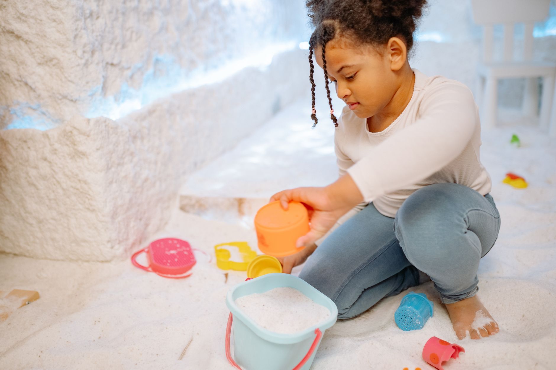A child playing with colorful toys in a sandy playroom, engaging with a bucket.