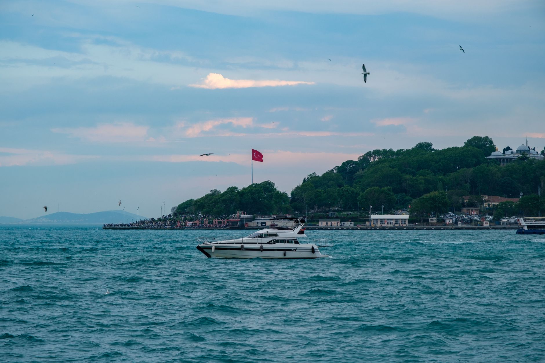 A tranquil Bosphorus view at sunset with a boat, Turkish flag, and green hills.