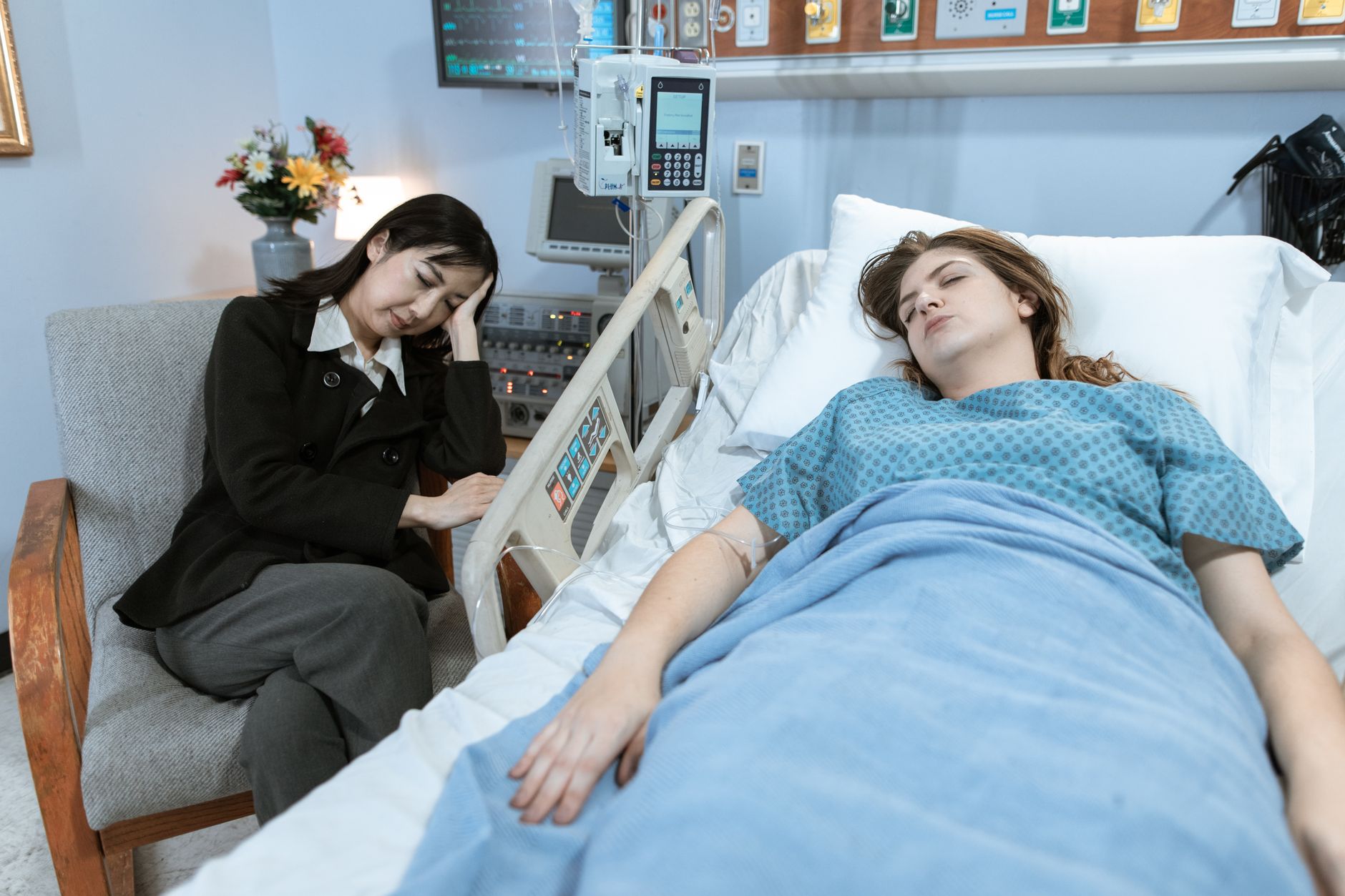 A woman sits beside a patient in a hospital bed, both looking distressed.