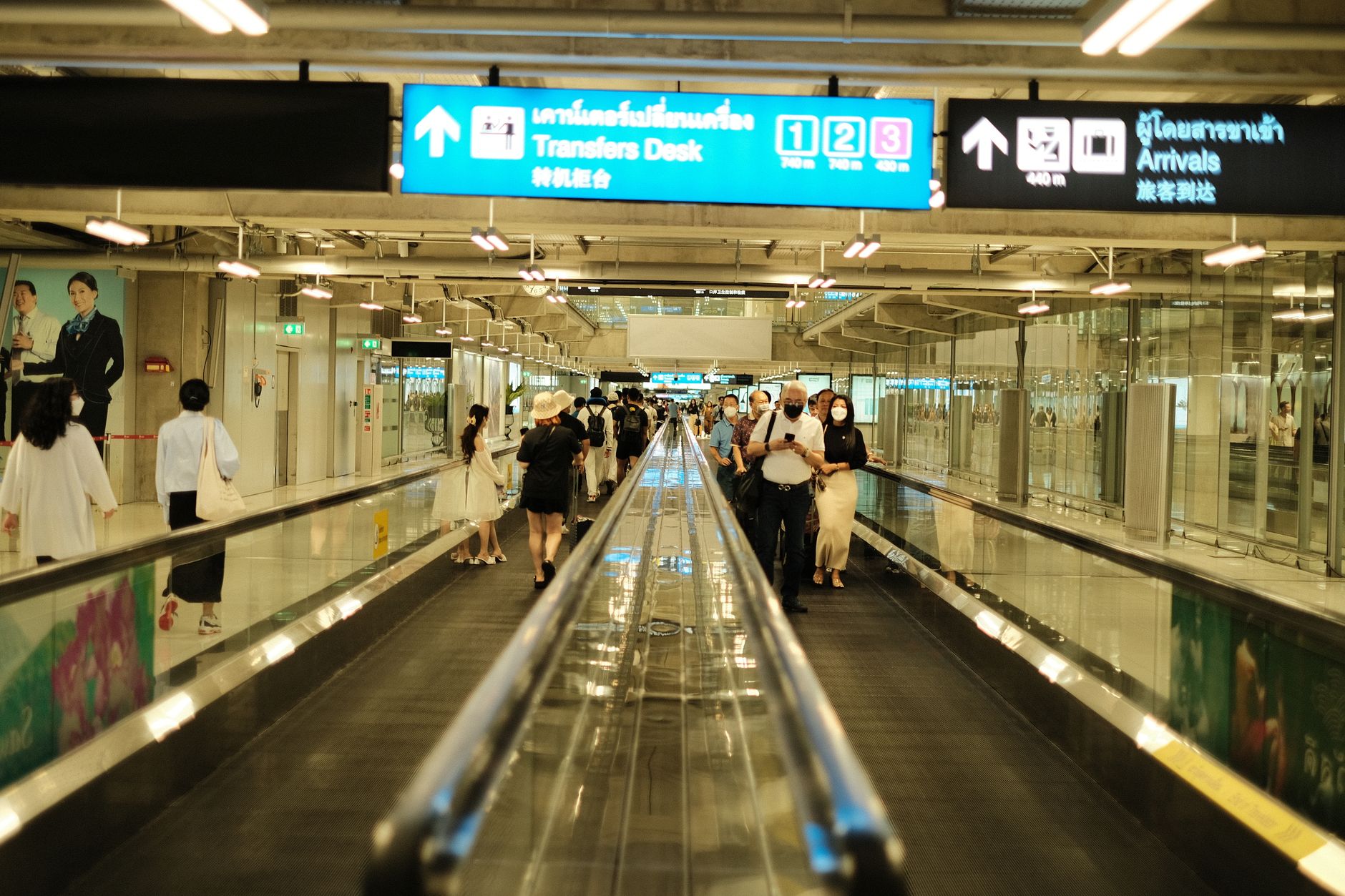 Passengers walking on a moving walkway toward the Transfers Desk at Bangkok Airport.