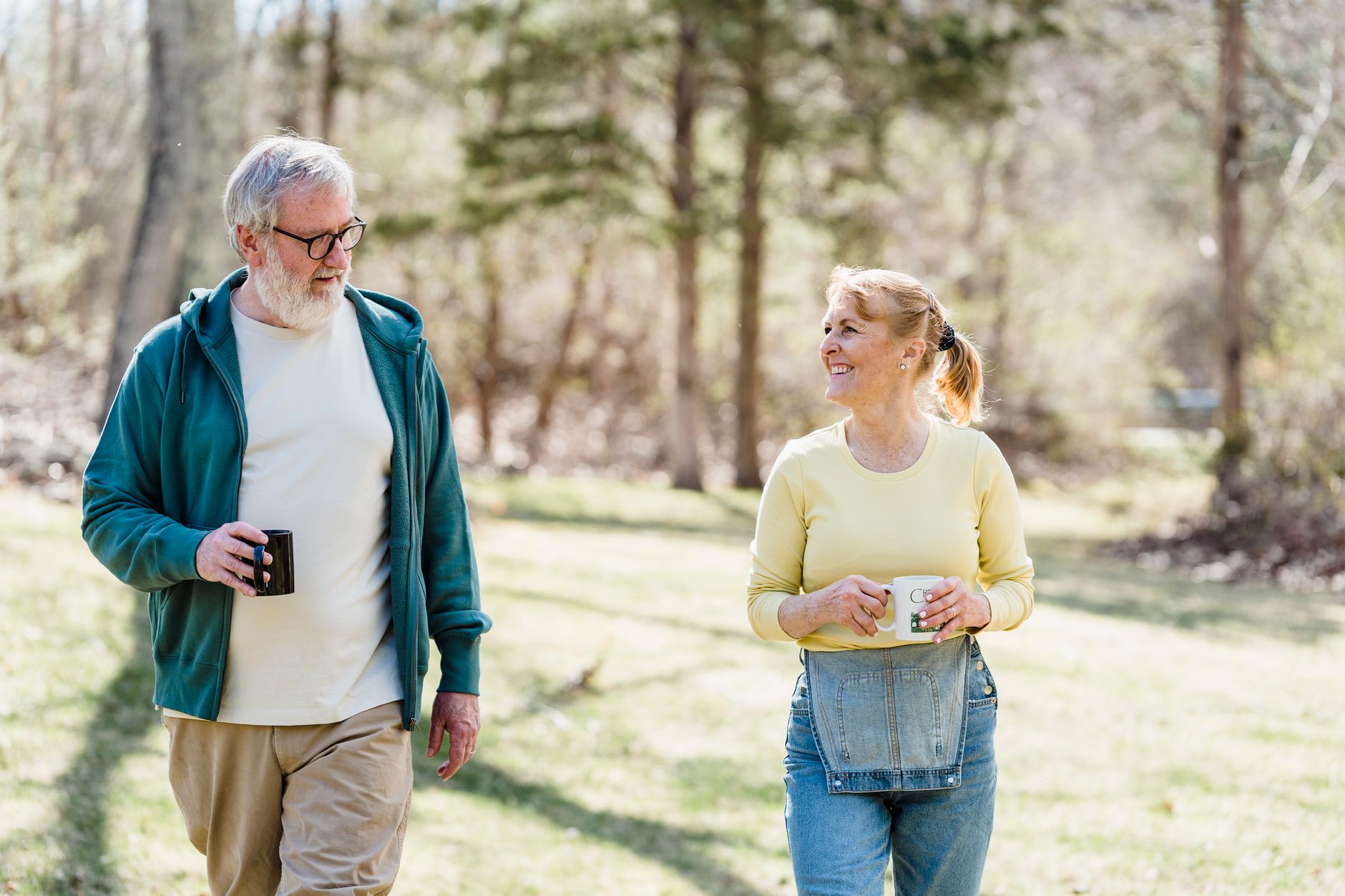 A senior couple walking in a park, smiling and holding coffee mugs, enjoying their time together.