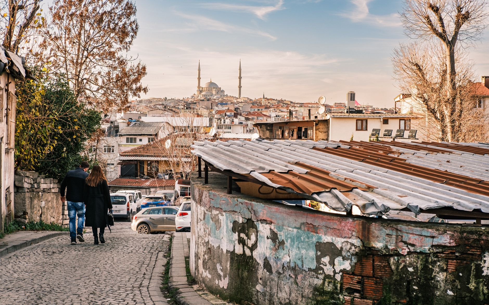 A couple walks hand in hand along a cobblestone street in Istanbul, with historic buildings and minarets in the background.