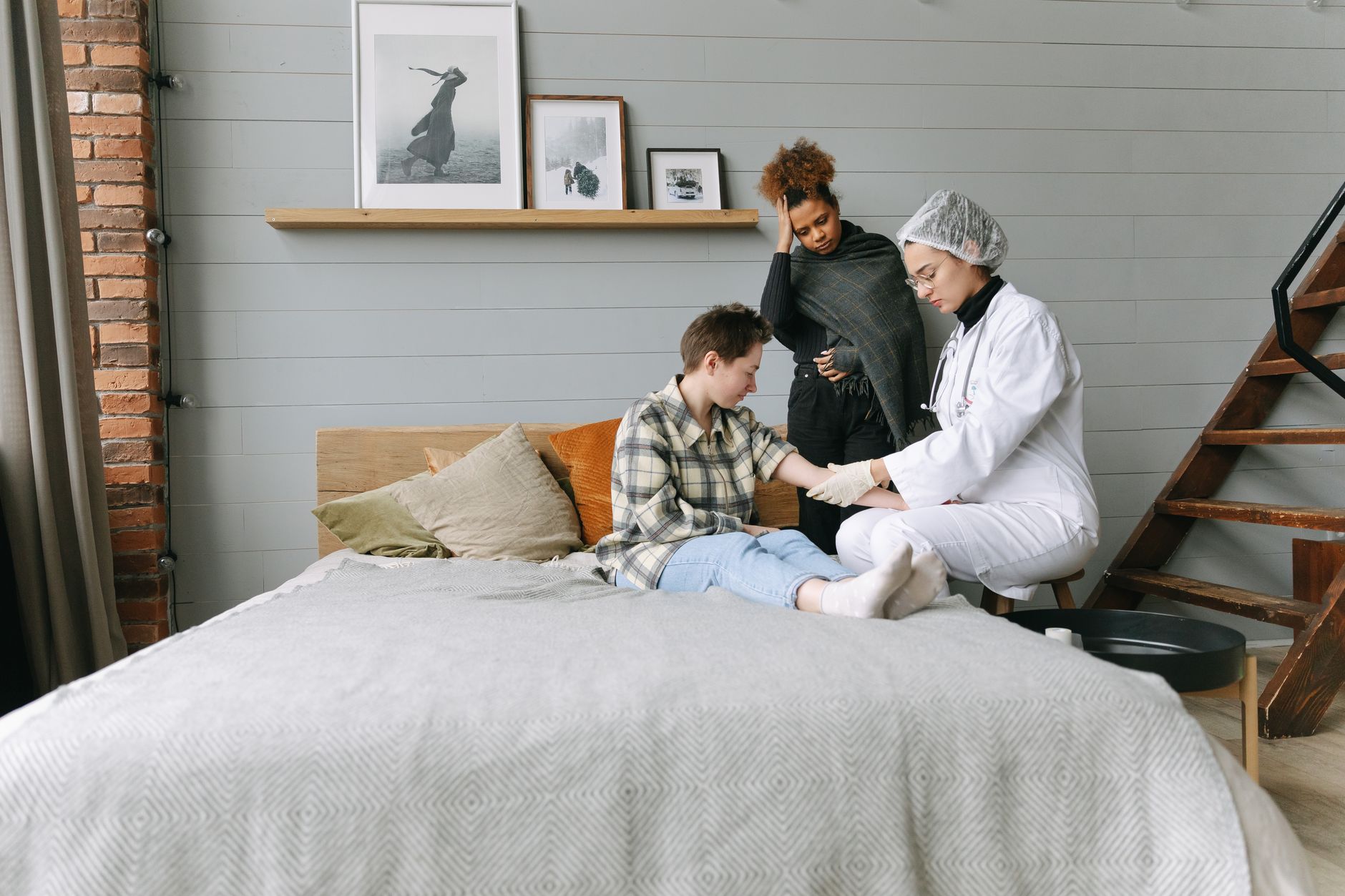A patient at home receiving care from a healthcare professional while being comforted by a friend.
