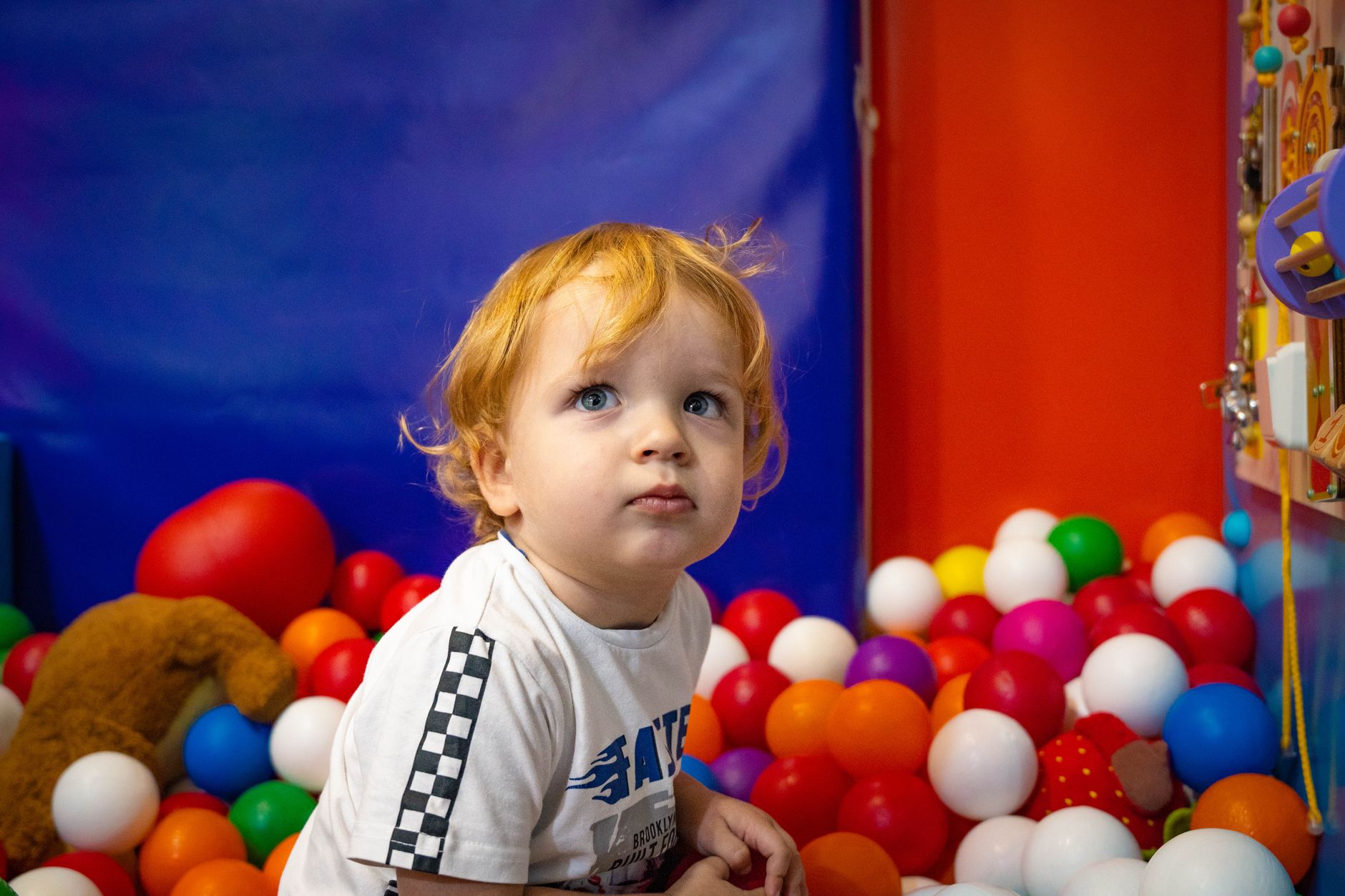 A child with curly red hair plays in a colorful ball pit in a hospital's playroom.