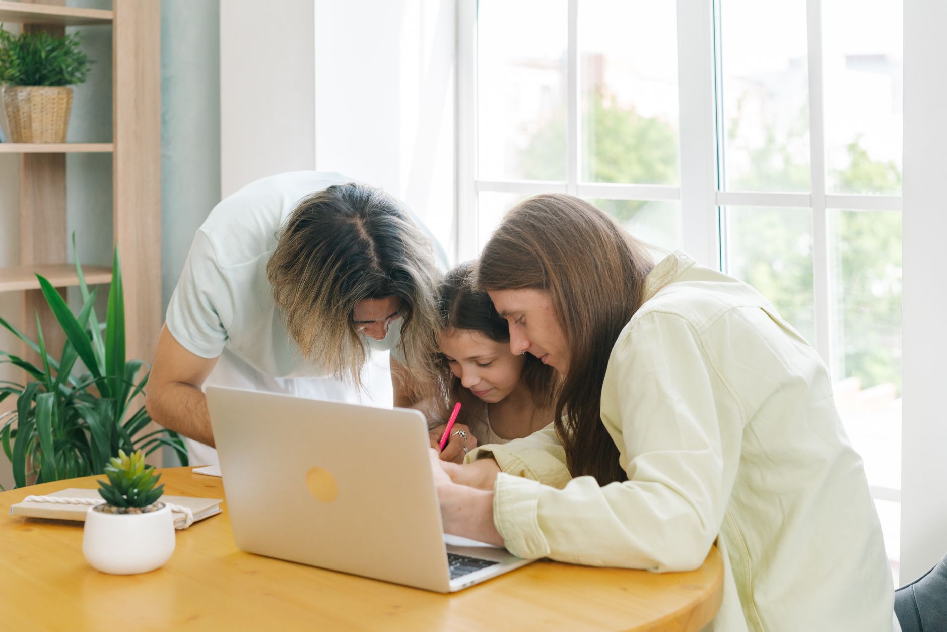 A family engages around a laptop, helping a young girl with learning tasks at home.