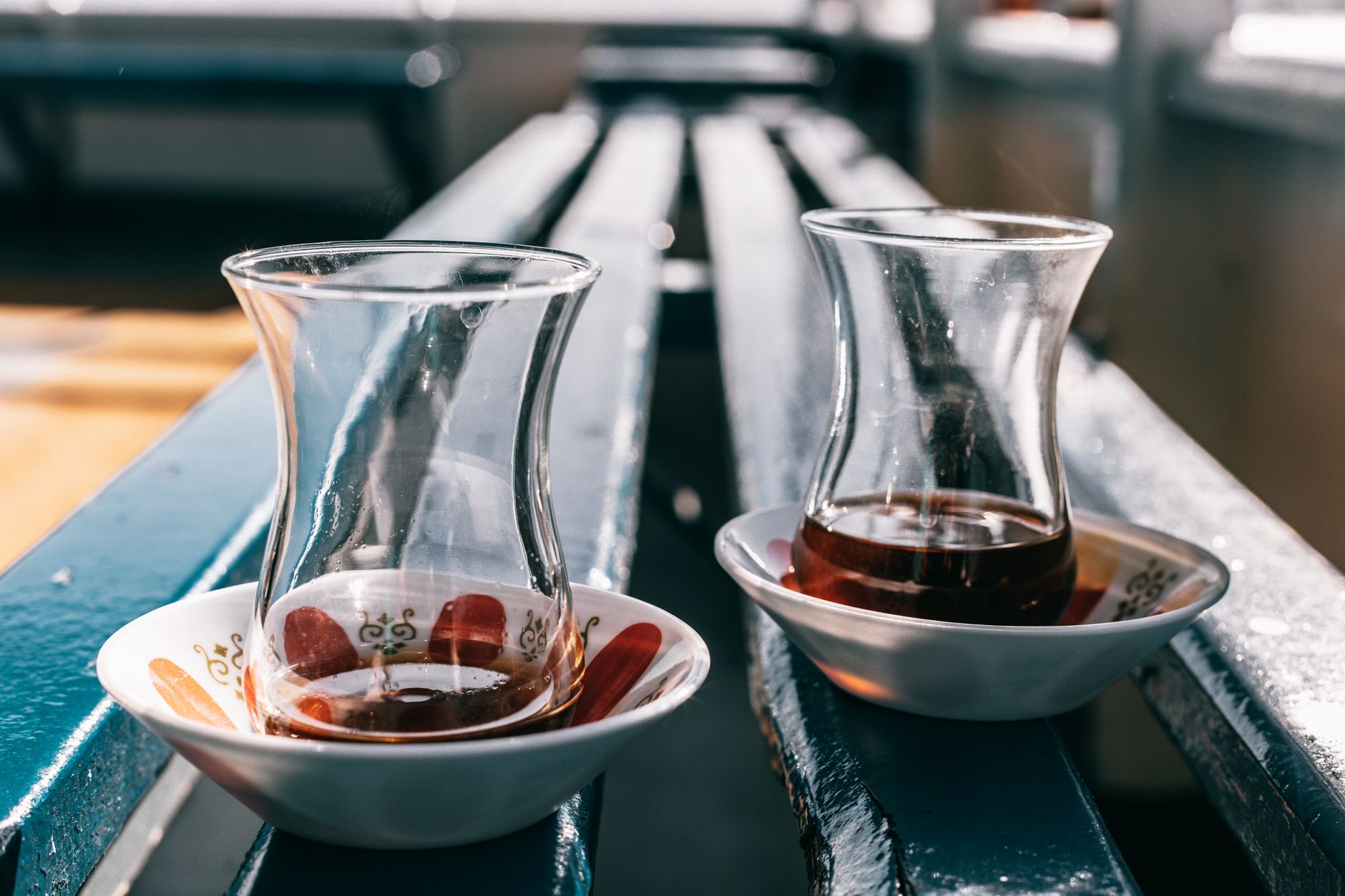 Two elegant glass tea cups on saucers, set on a blue bench near water.