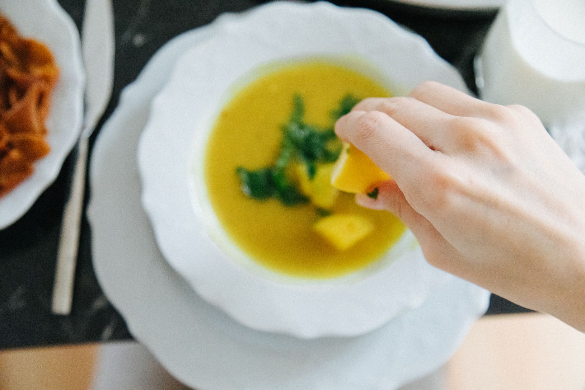 A hand adds lemon to a bowl of Turkish lentil soup, featuring vibrant greens and a side of bread.