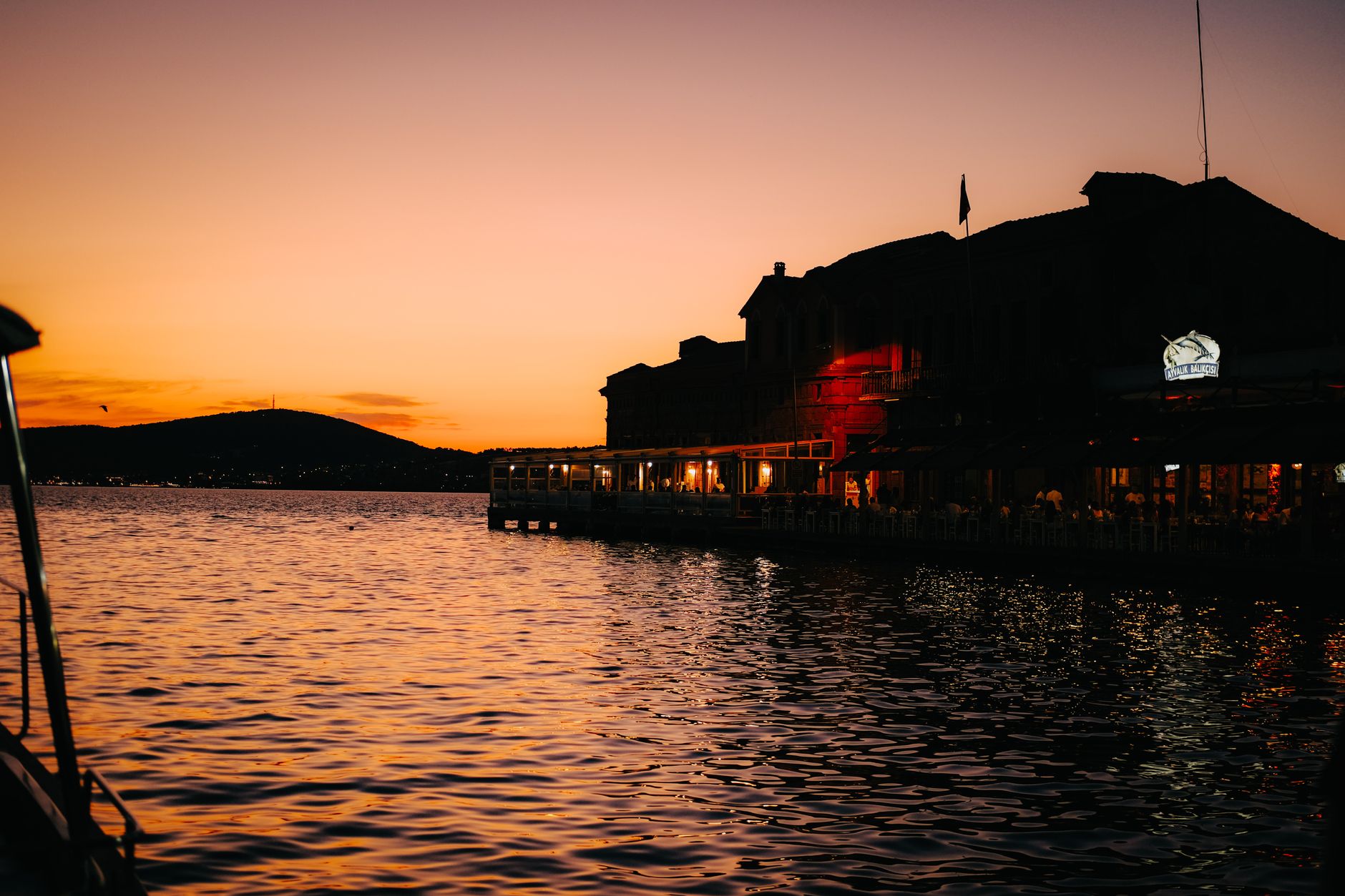 A serene sunset over the water in Kaş, Türkiye, with silhouettes of buildings and gentle waves.