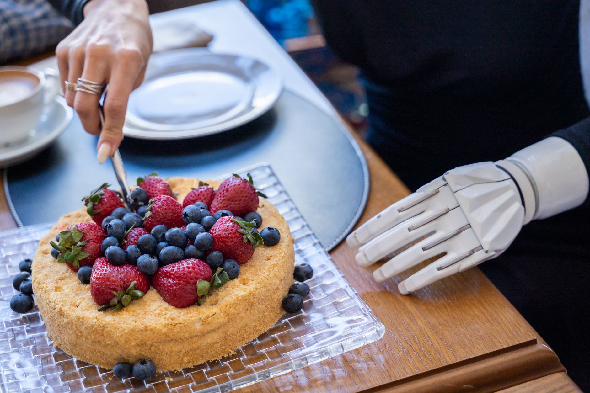 A patient uses a robotic exoskeleton hand to slice a cake topped with strawberries and blueberries.