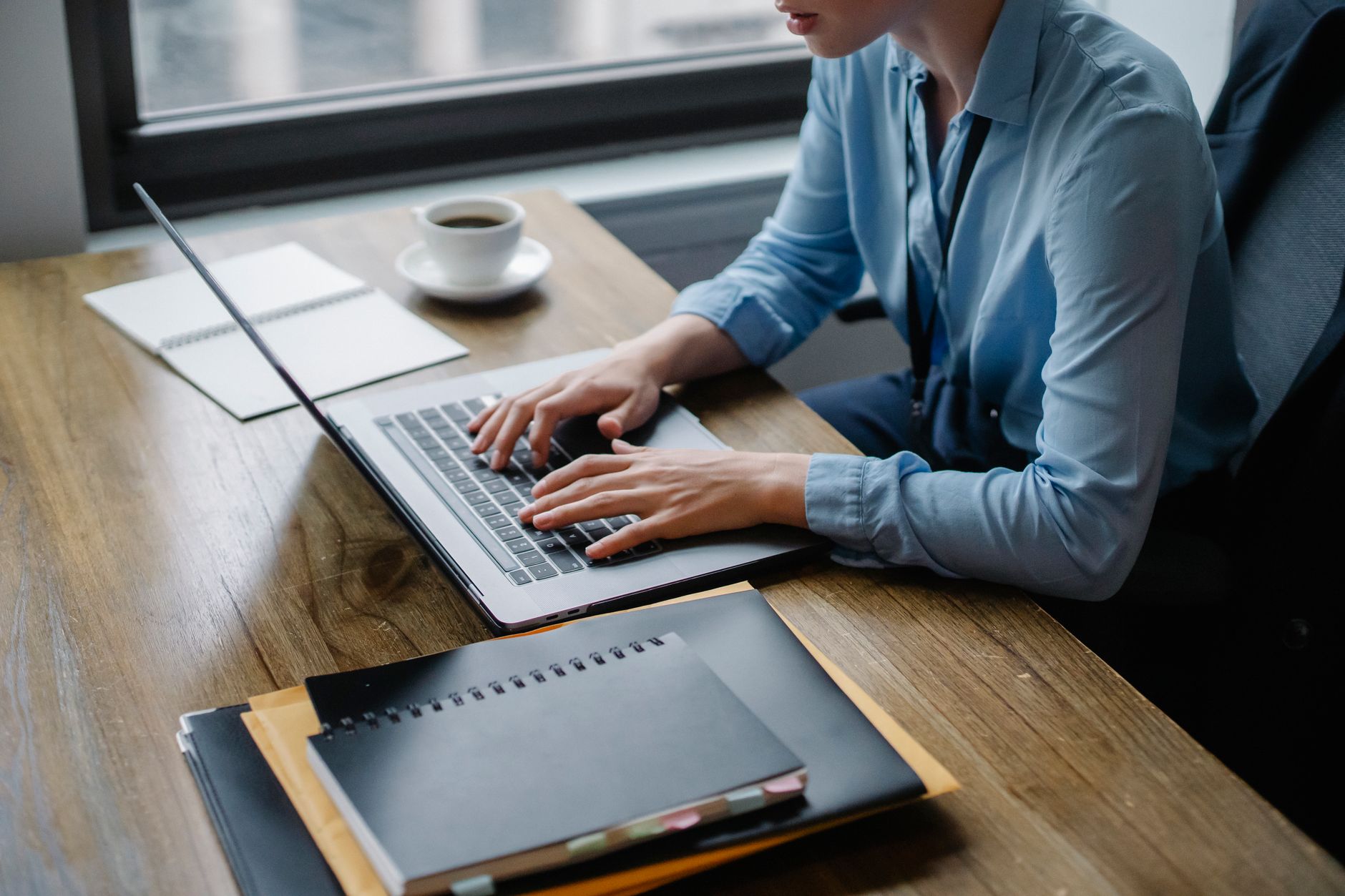 A person working on a laptop with a cup of coffee, notebooks on a wooden desk.