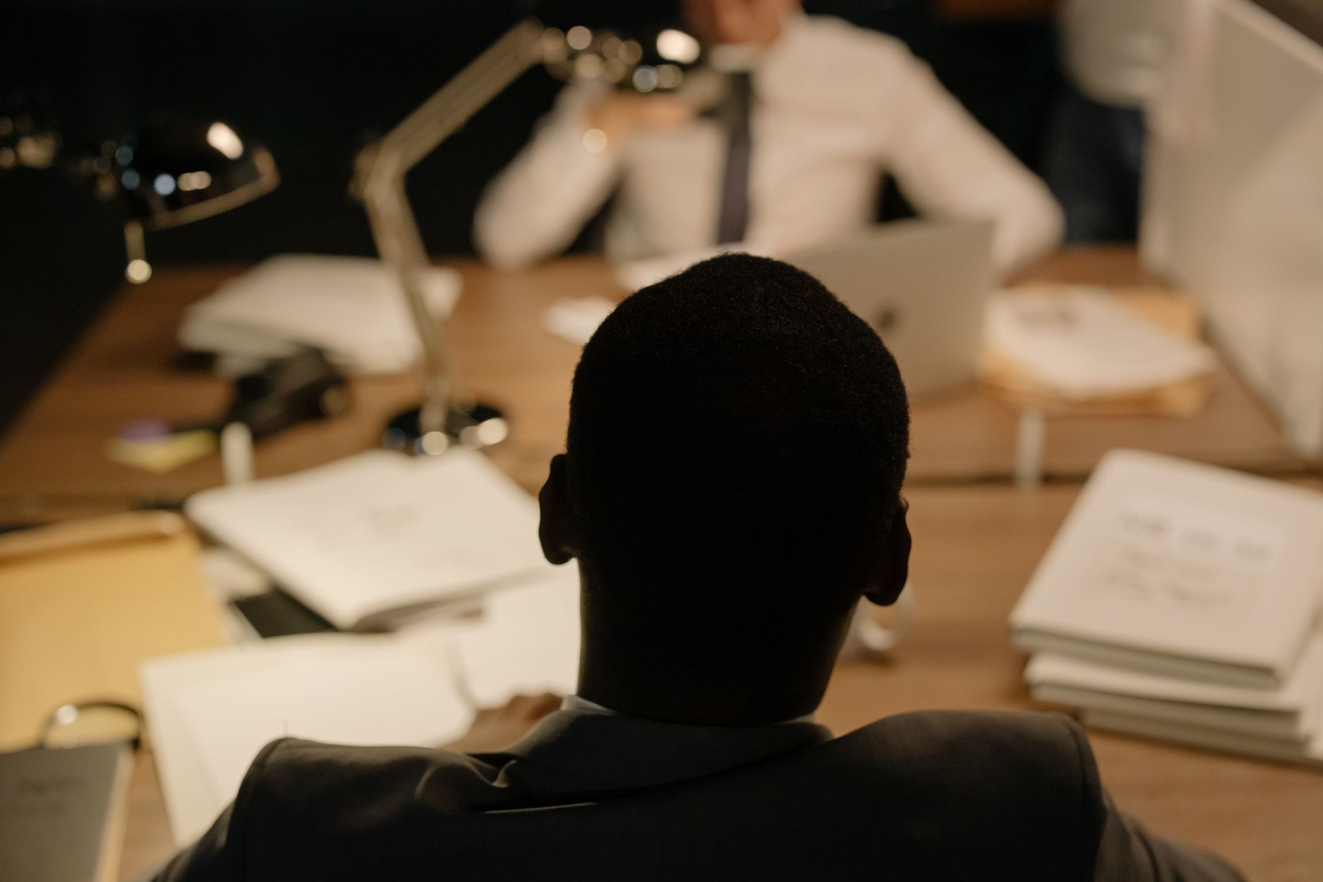 A man in a suit sits confidently at a desk in an office, engaging in a conversation.
