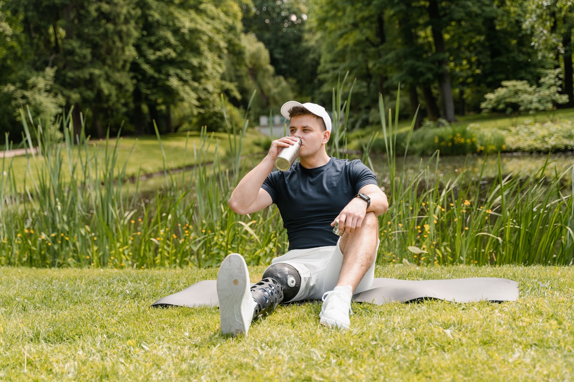A young man with an artificial leg sits on a mat in a serene park, sipping a drink amidst lush greenery.