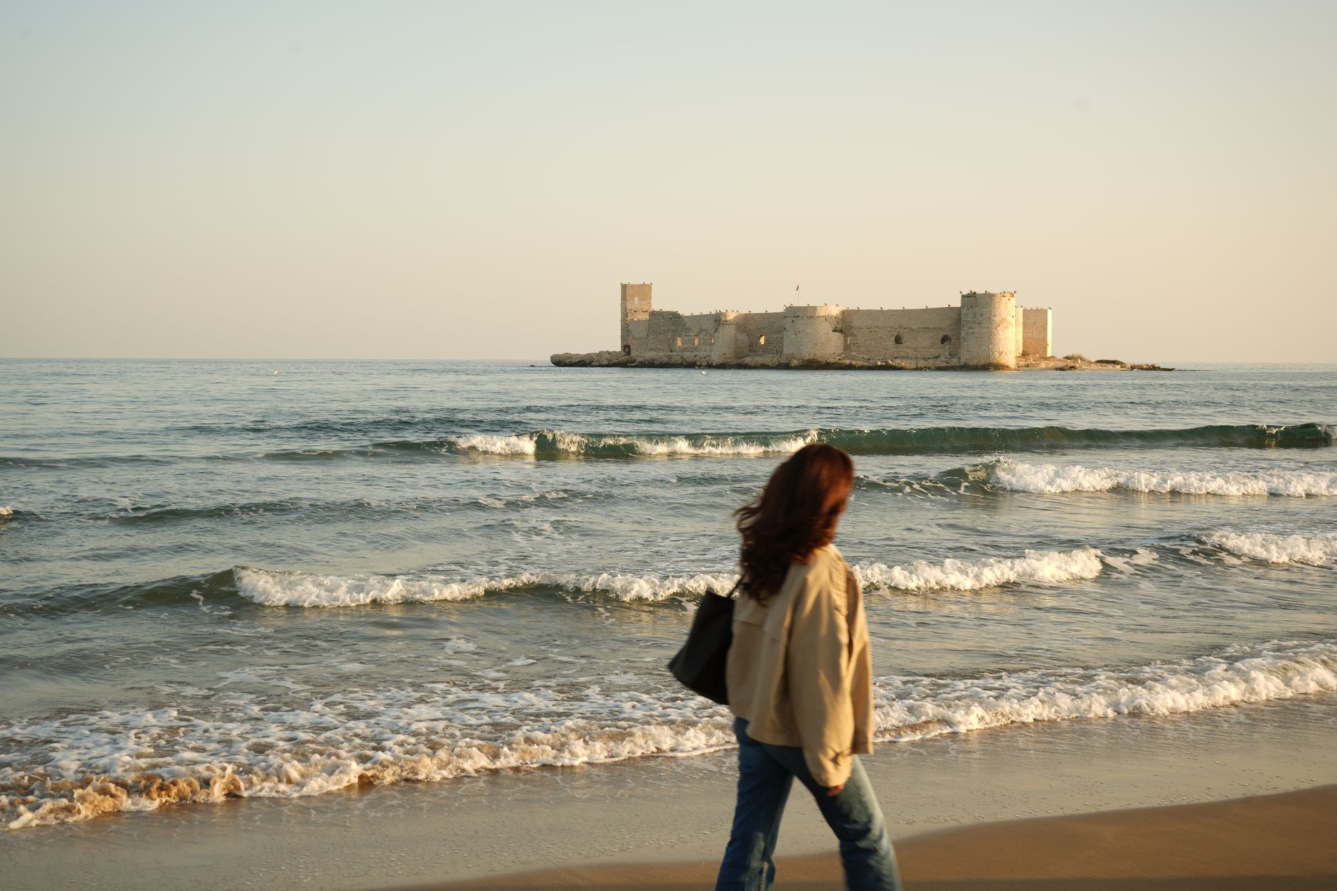 A woman strolls along the beach near Maiden's Castle in Mersin, with gentle waves lapping at the shore.