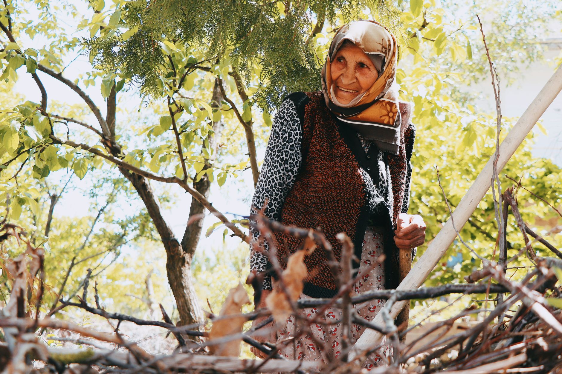 An elderly woman with a joyful smile stands amidst trees in a lush garden.
