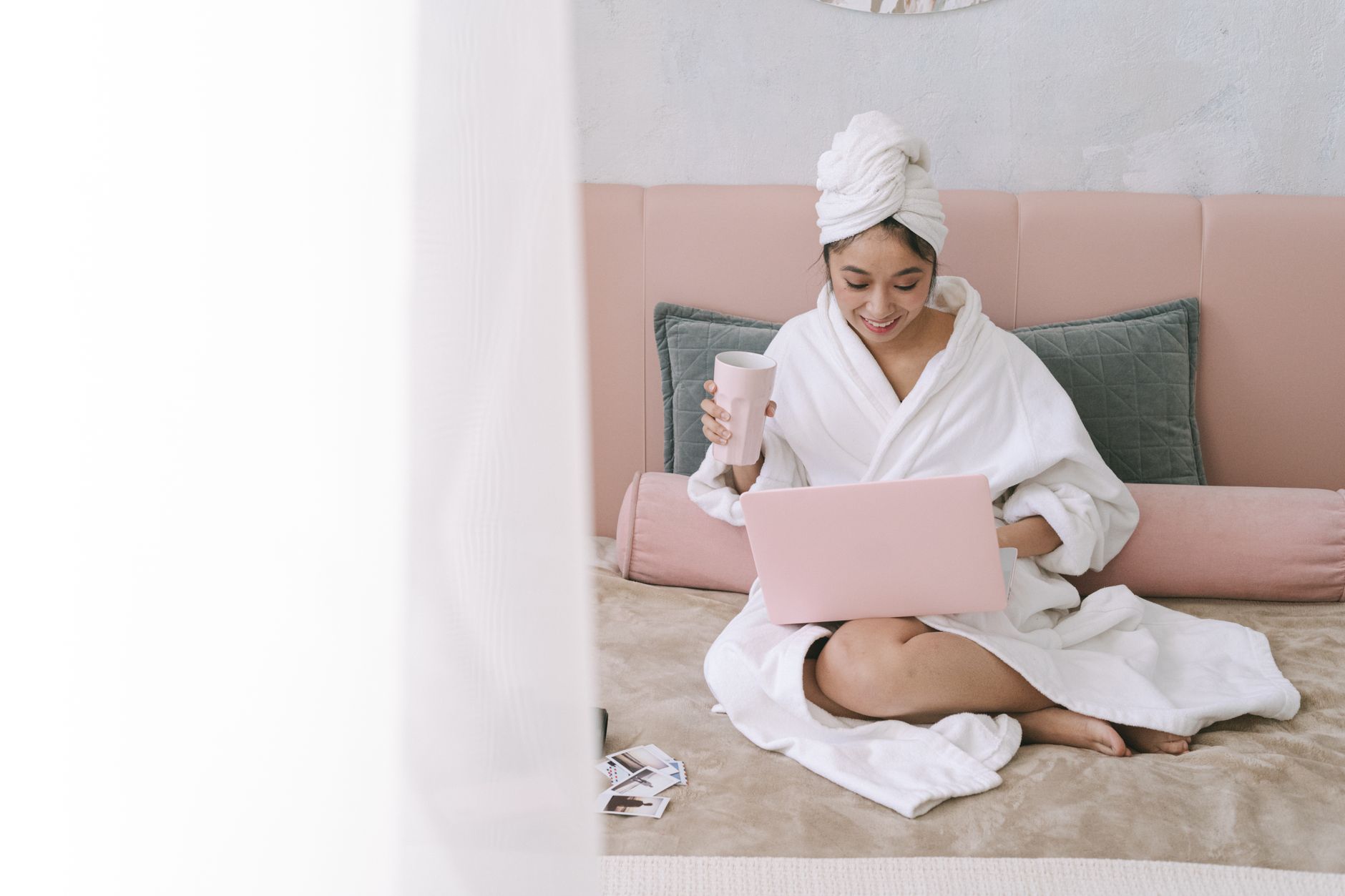 A woman in a white robe sits on a bed with a laptop, enjoying a drink in a calming home environment.