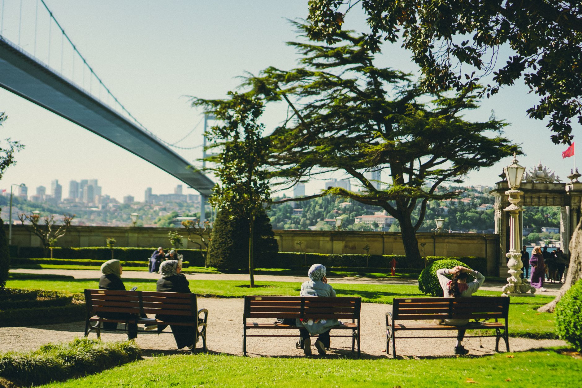 People sitting on benches in a garden overlooking Istanbul's skyline and Bosphorus Bridge.