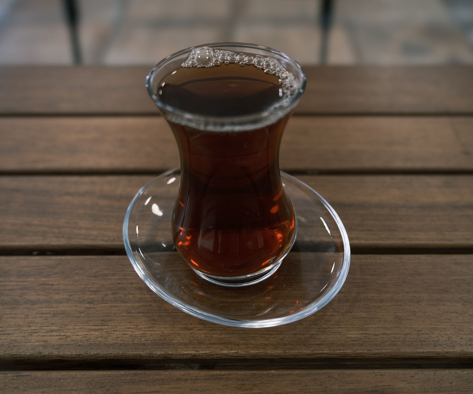 A glass of tea with bubbles sits on a clear saucer on a wooden table.