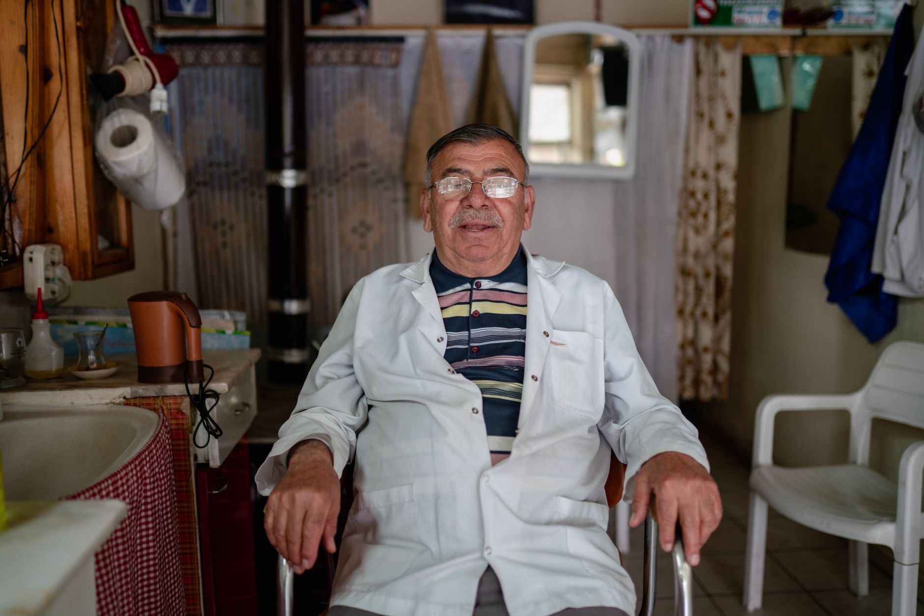 An elderly man wearing glasses and a white coat smiles while sitting in a modern hospital setting.