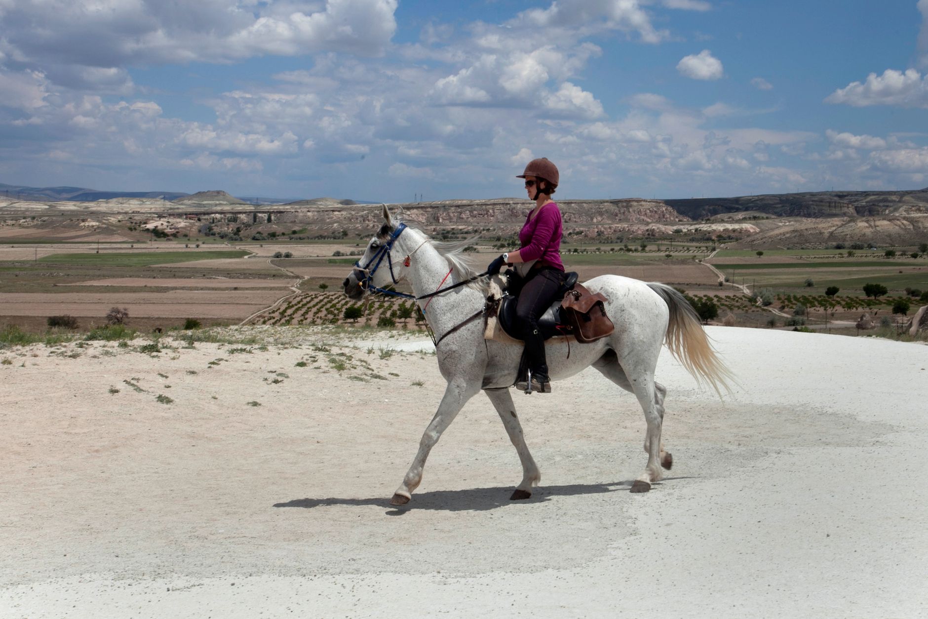 A rider in a purple top riding a white horse across the green Zara Plain with hills in the background.