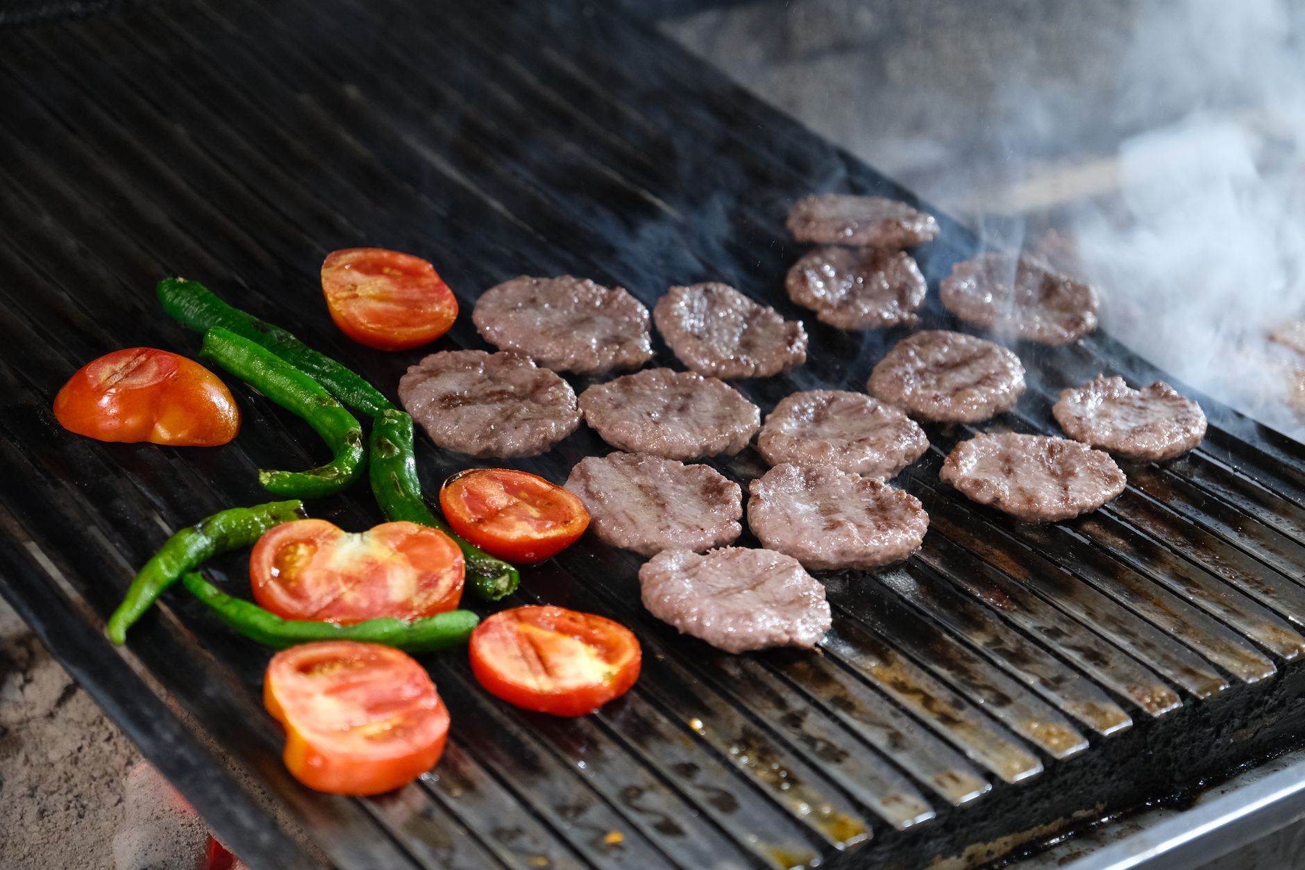 Sivas köfte grilling on a barbecue with tomatoes and green peppers.