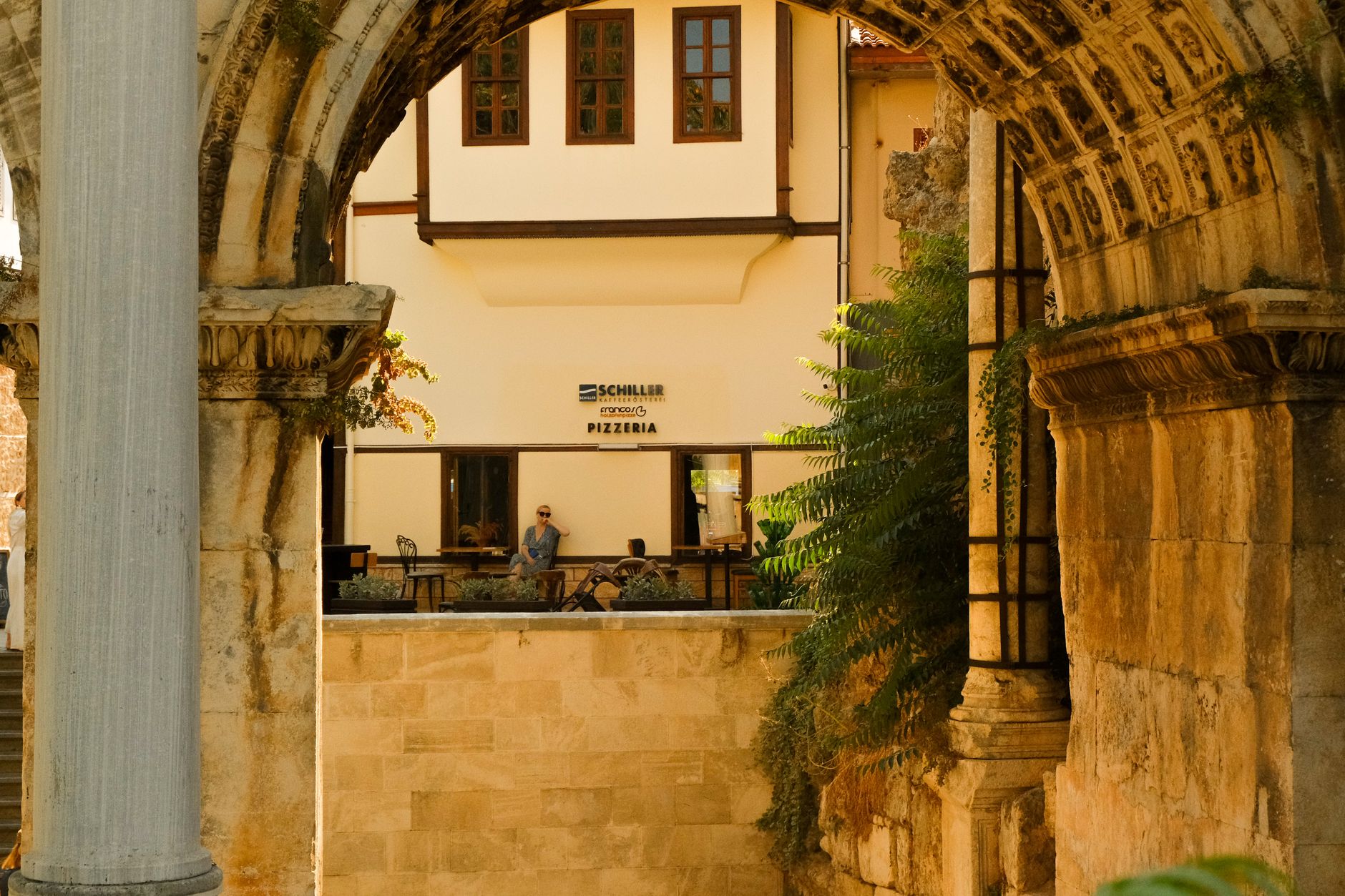 A man relaxes by a pizzeria near Hadrian's Gate, framed by ancient architecture.