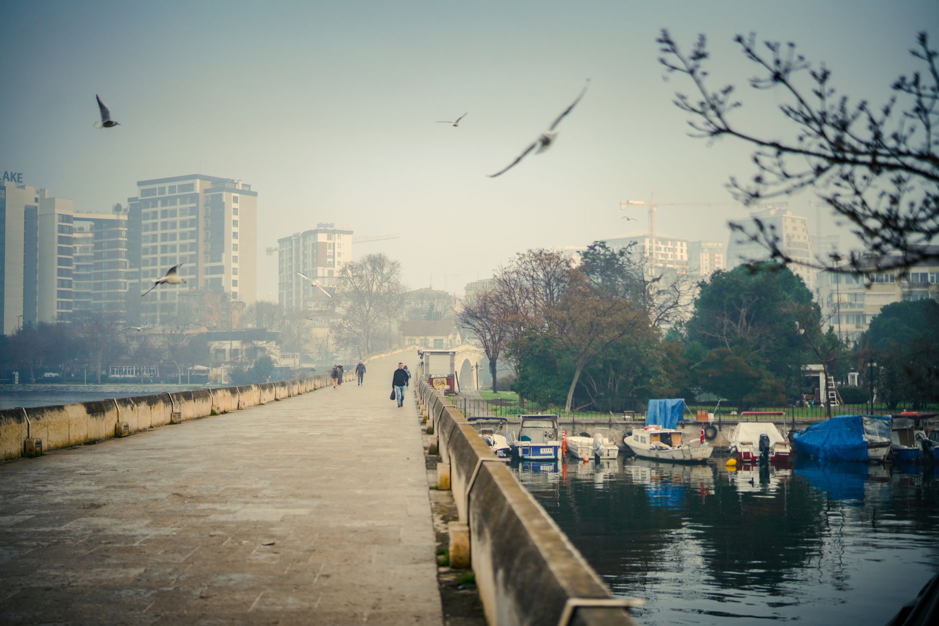 People walking along the Porsuk River promenade with boats and buildings in the background.