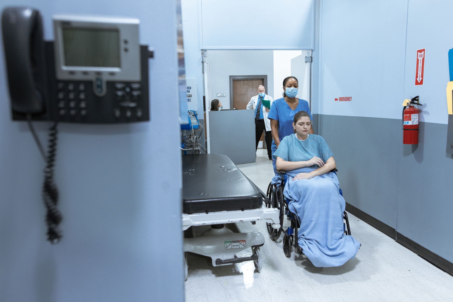 A woman in a wheelchair is escorted by a nurse in scrubs through a hospital hallway after surgery.