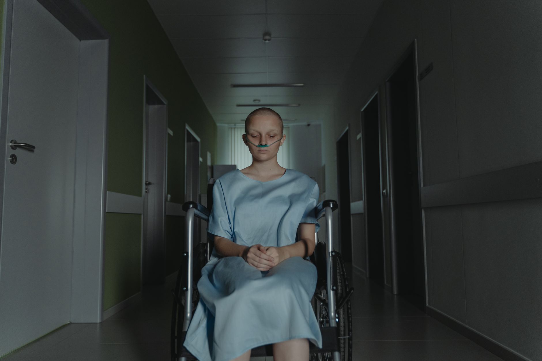 A bald patient sits in a wheelchair in a hospital corridor, wearing a blue gown.