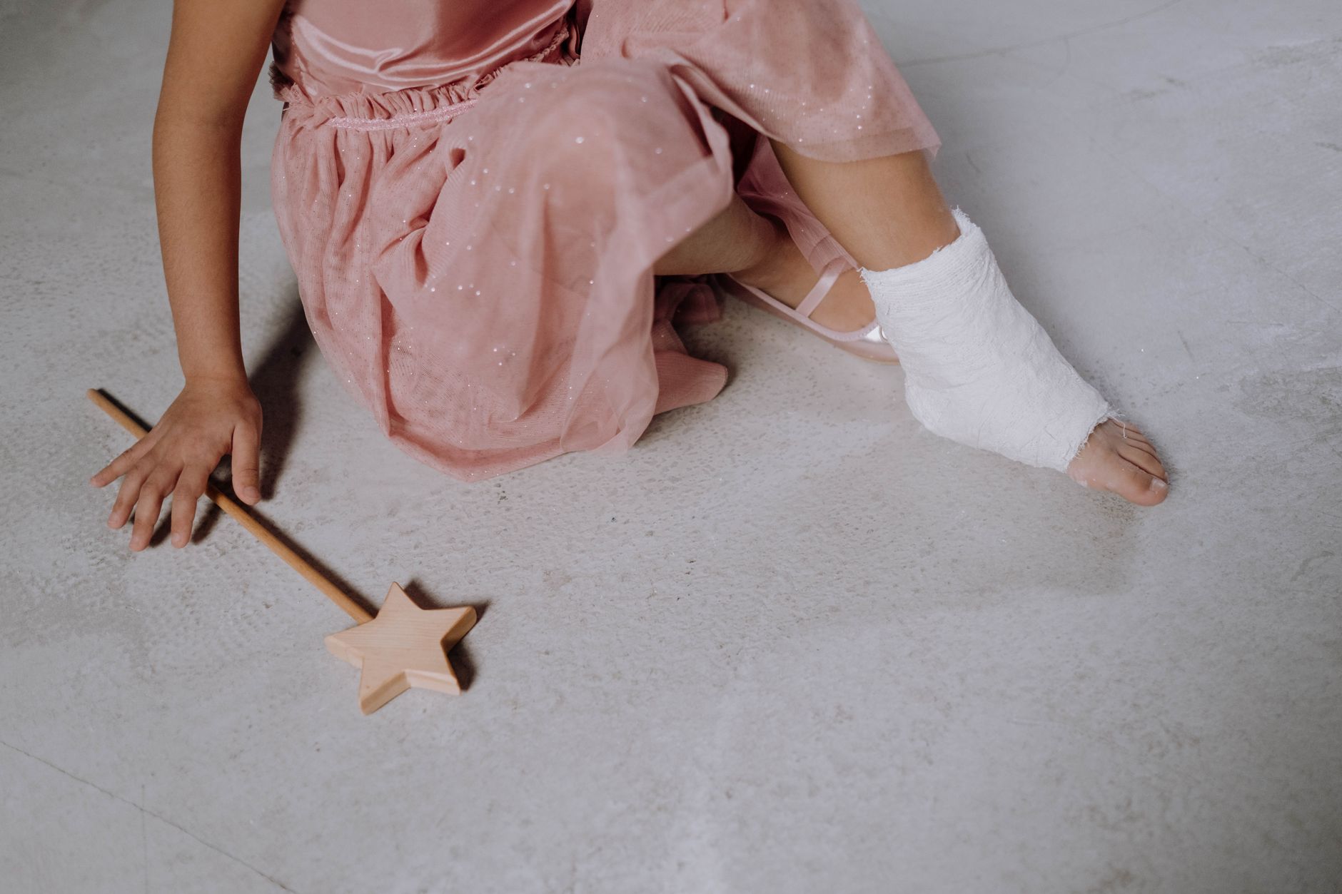 A child in a pink dress with a bandaged foot sits on a floor with a wooden star wand.
