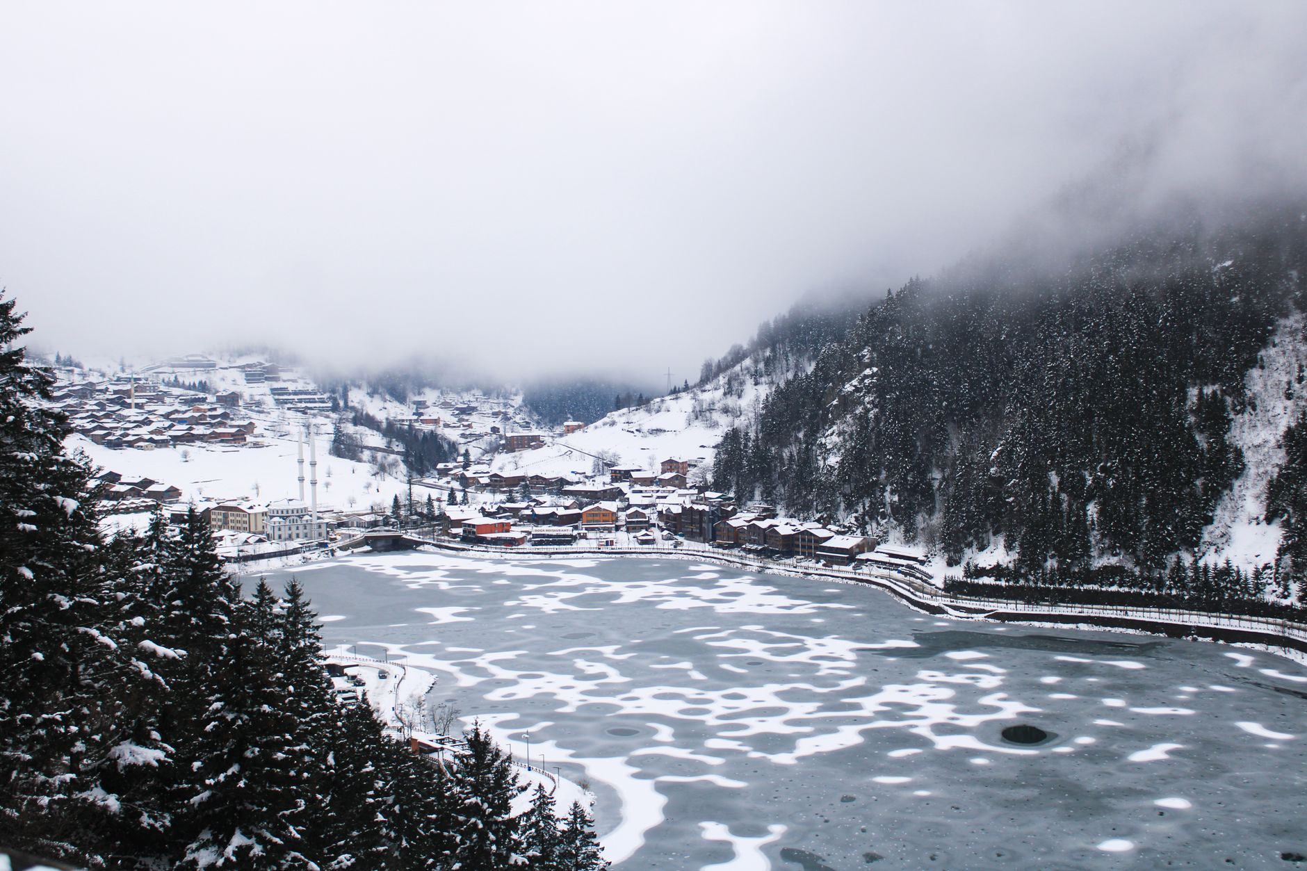A tranquil snowy view of Trabzon with a frozen lake, surrounded by misty mountains and quaint houses.