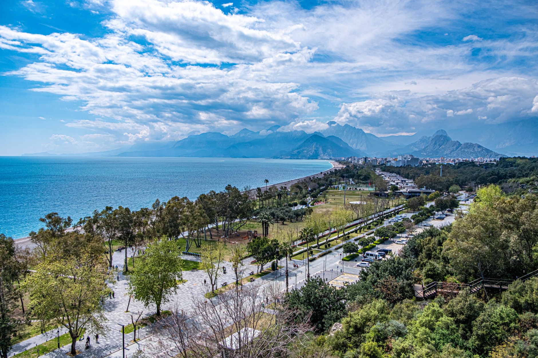 A scenic view of Antalya's seaside promenade, featuring lush greenery and mountains under a bright blue sky.