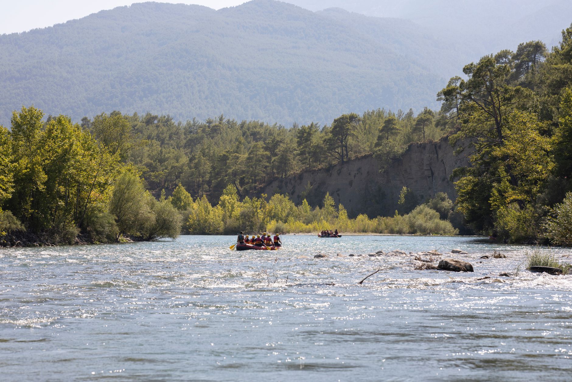 Rafting yapanlar, yemyeşil doğa ve dağlar arasında berrak bir nehirde akıntılarda ilerliyor.