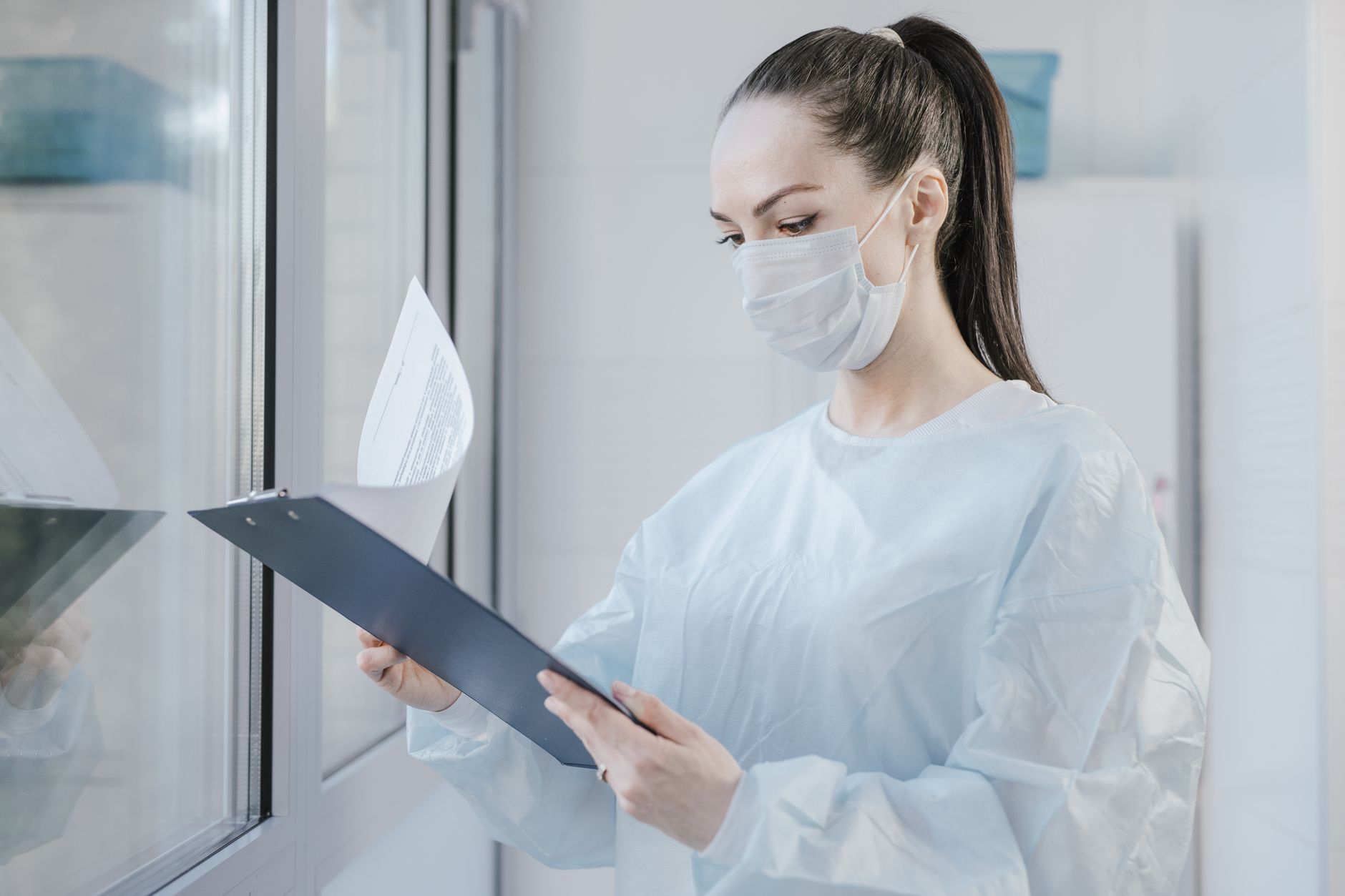 Healthcare professional in mask examining paperwork by window.