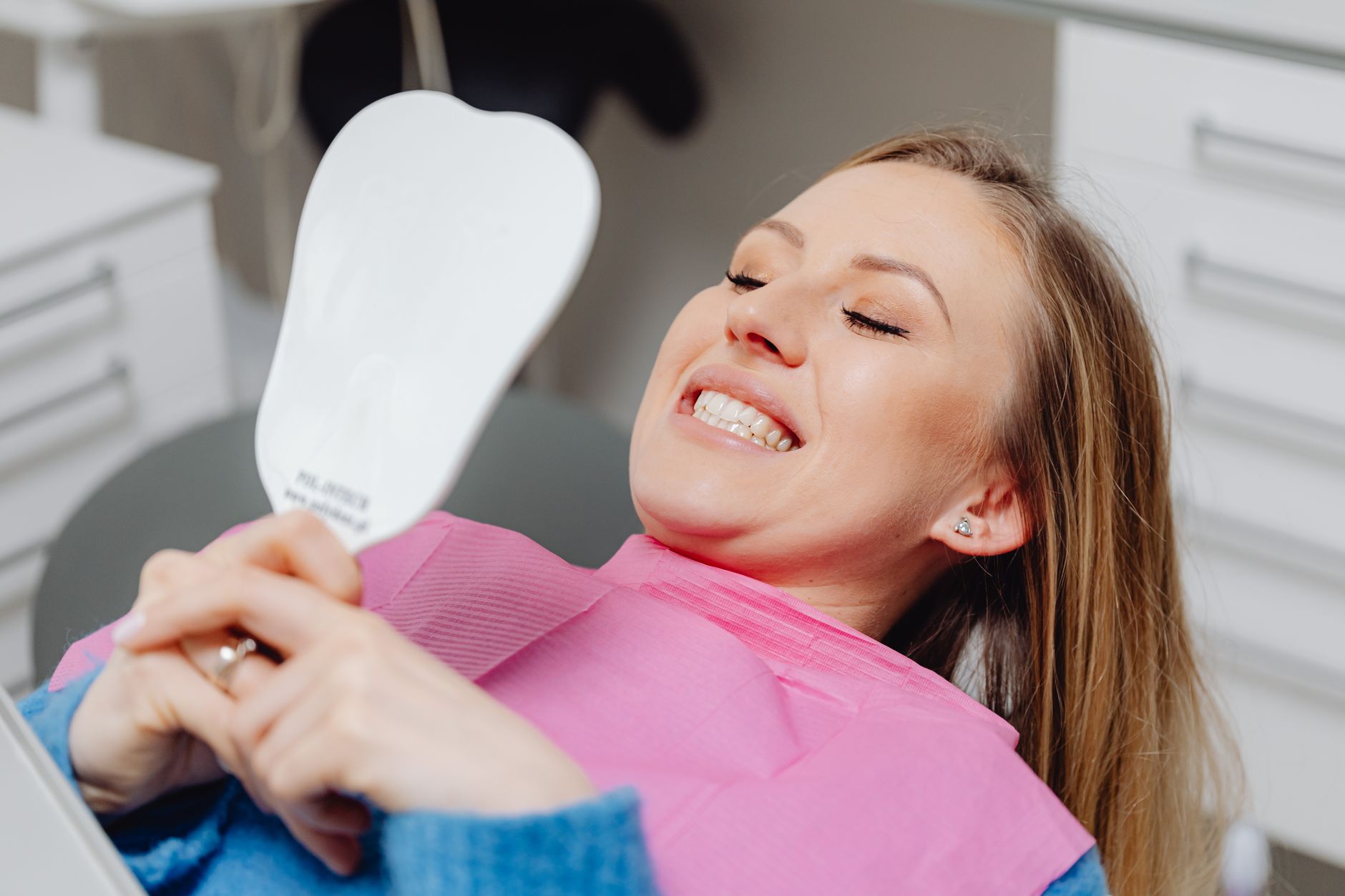 A woman with a confident smile holds a mirror, showing her healthy teeth after dental treatment.