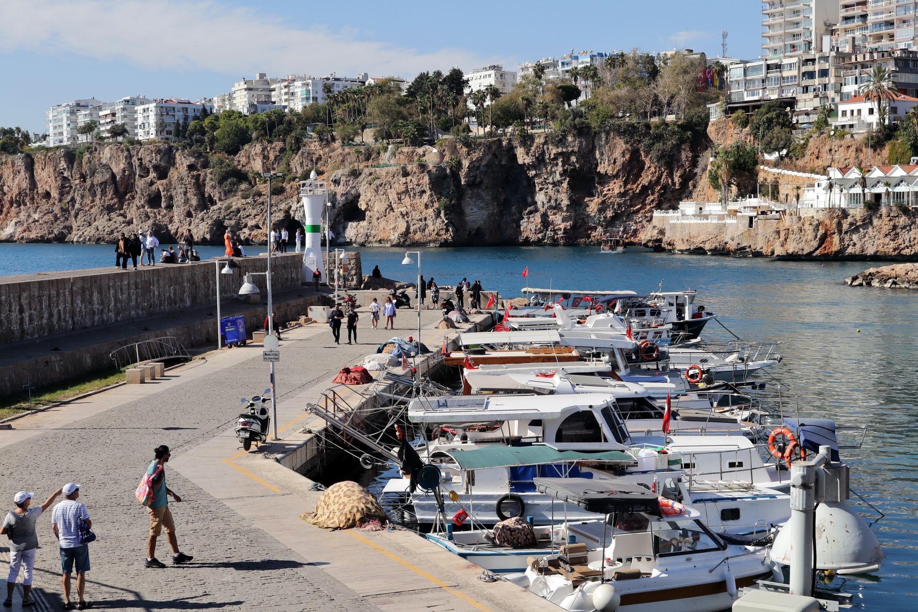 A bustling marina in Antalya with boats, visitors walking, and scenic cliffs.
