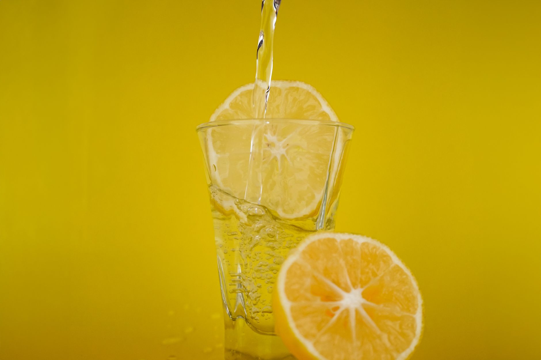 A glass of water with lemon slices against a bright yellow background.