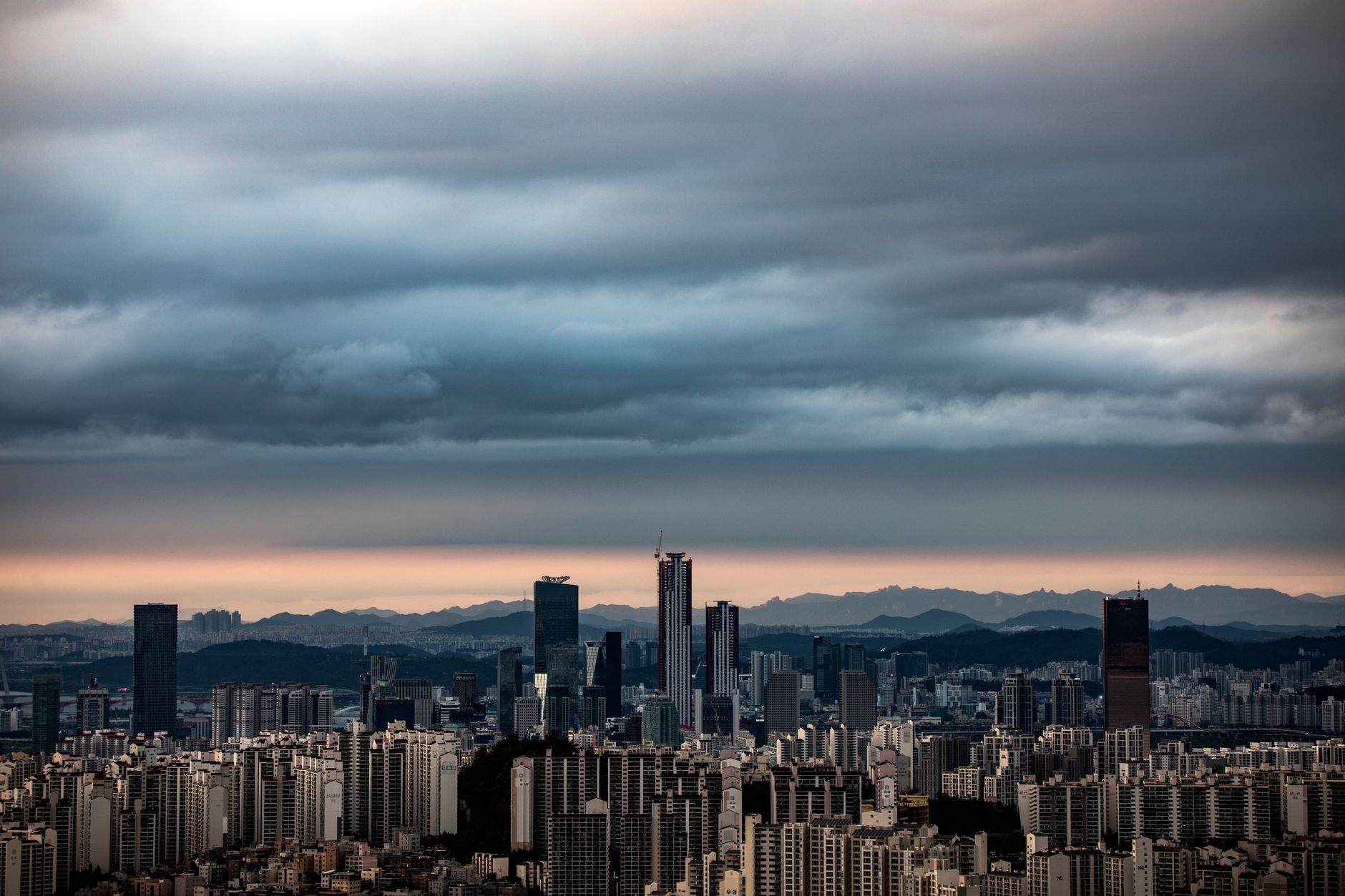 A panoramic view of Seoul's Gangnam district skyline with dark clouds, showcasing high-rise buildings and distant mountains.