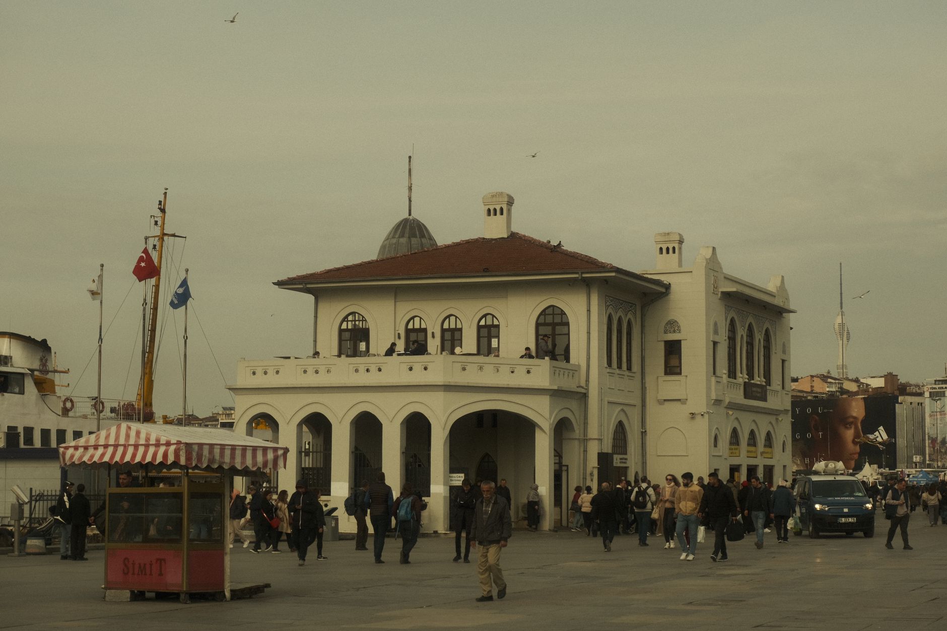 کنار آب کادی‌کوی شلوغ با مردمی که قدم می‌زنند، یک دکه سیمیت و معماری تاریخی.