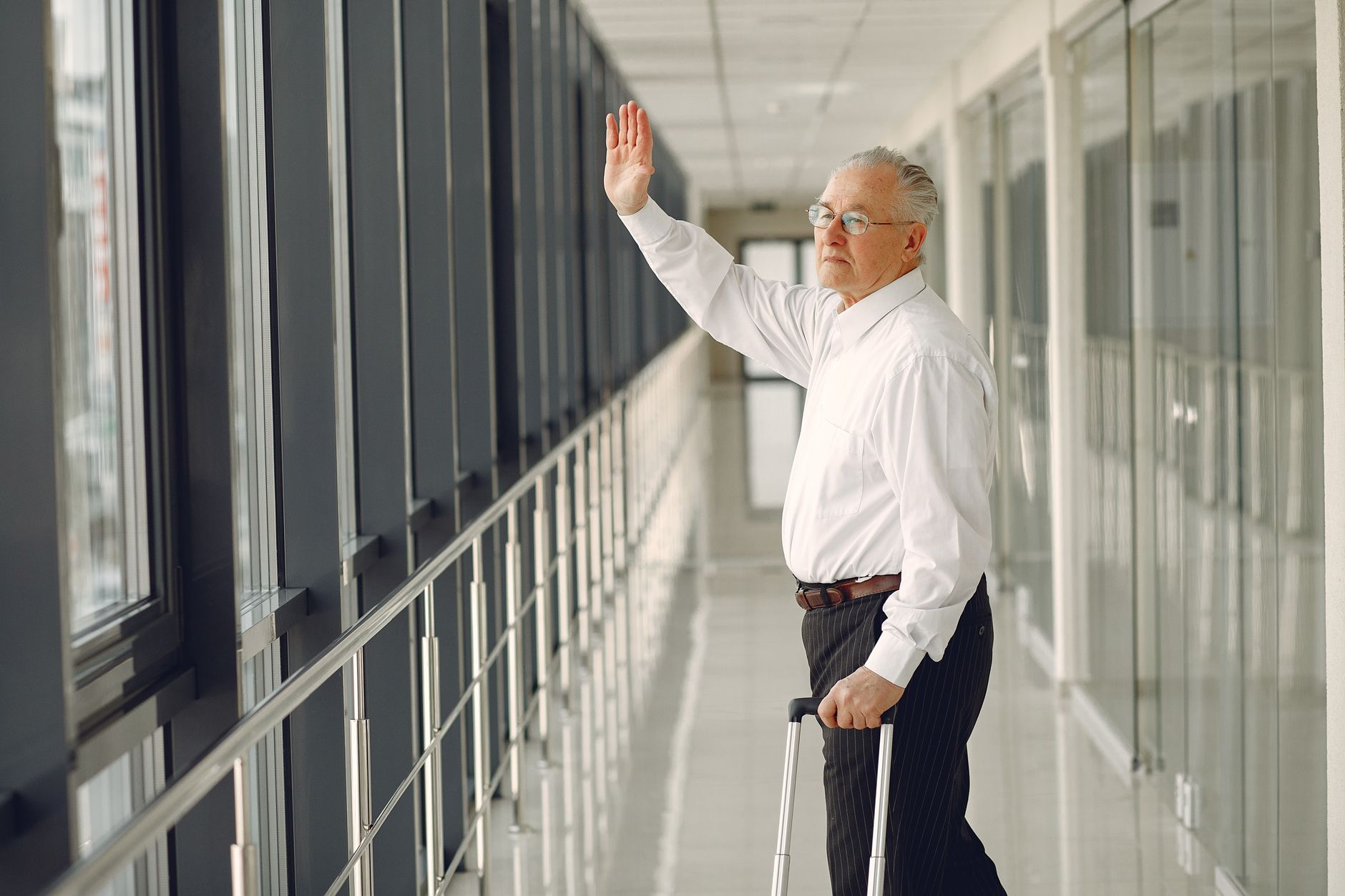 An elderly man in a white shirt waves goodbye while holding a suitcase in a sleek corridor.