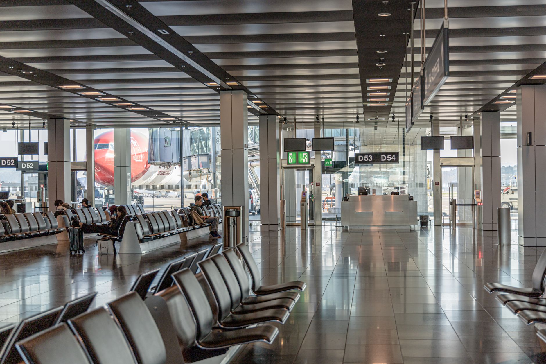 Spacious airport waiting area featuring flight gates D52 and D54, with travelers seated and a plane visible through the window.