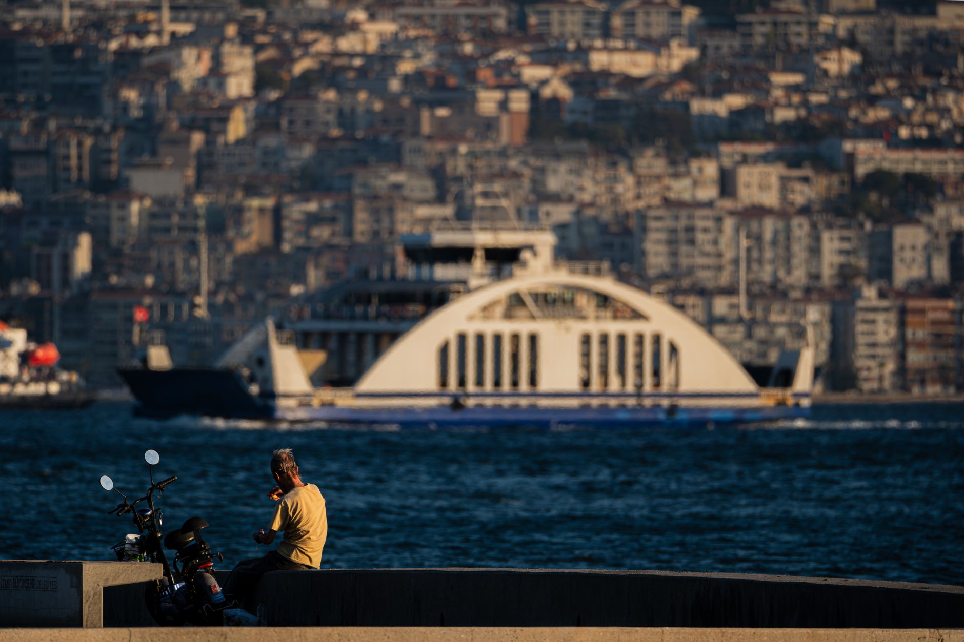 A person sitting by the Izmir waterfront, watching a ferry pass by amidst a cityscape.