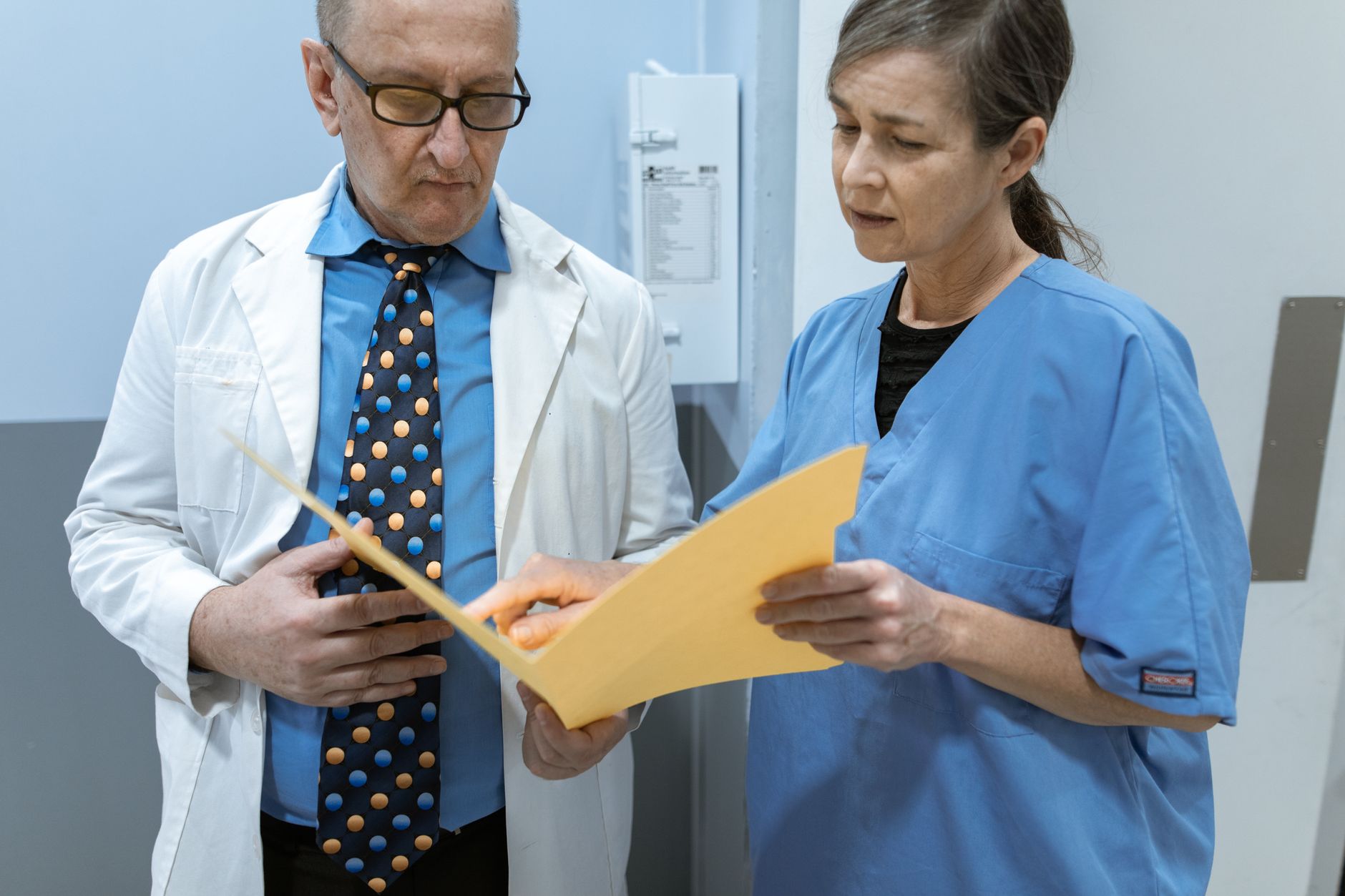 A doctor in a white coat and a nurse in scrubs discuss a medical summary in a clinic.