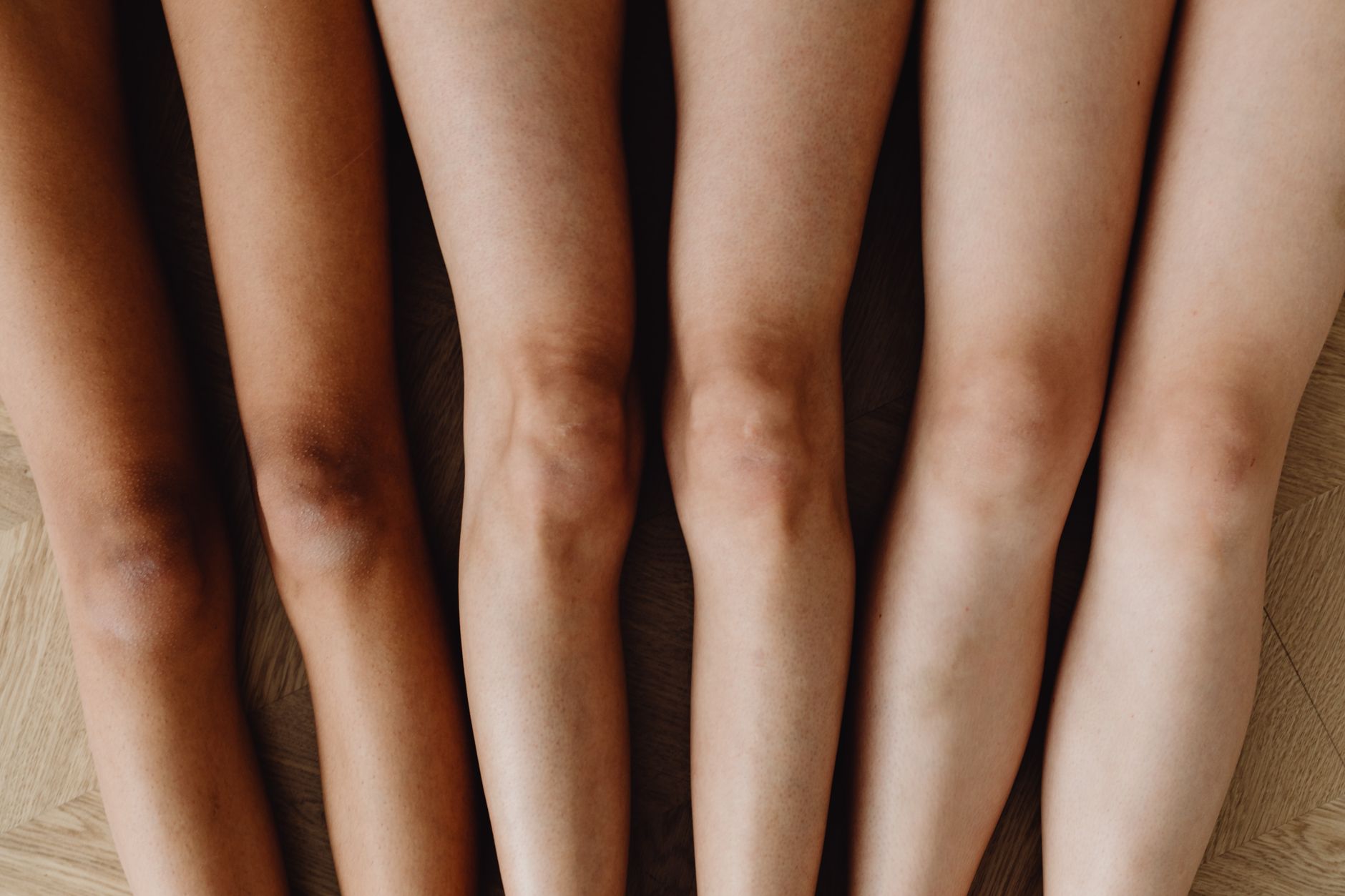 Five pairs of legs showing different skin tones lined up on a wooden floor.