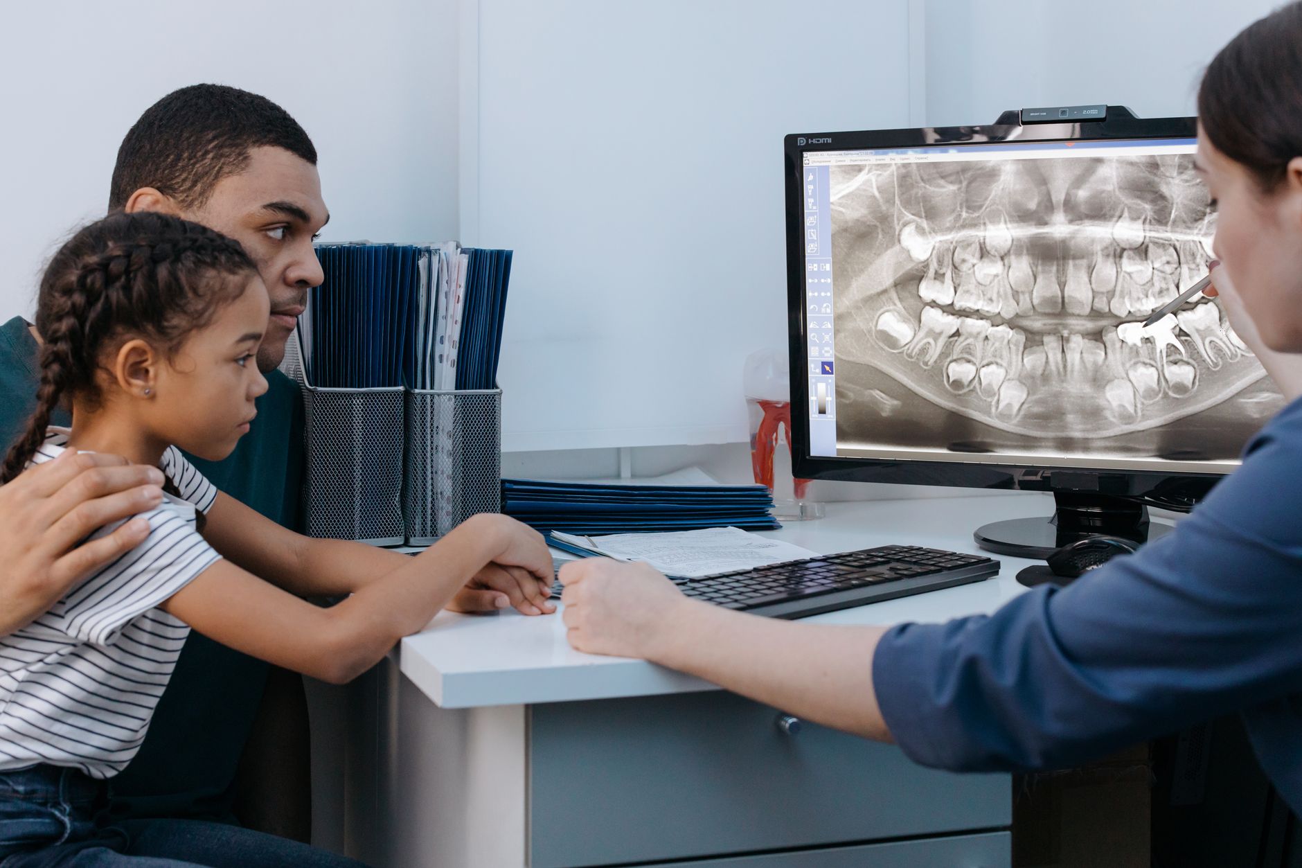 Pediatrician explains dental treatment to a father and daughter, showing an X-ray on computer.