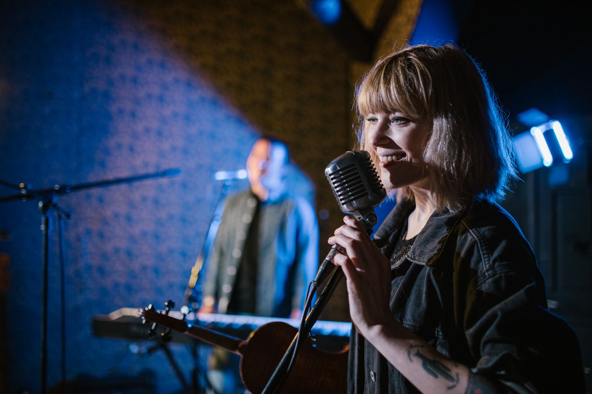 A female singer smiles while performing live, microphone in hand, with a male musician in the background.