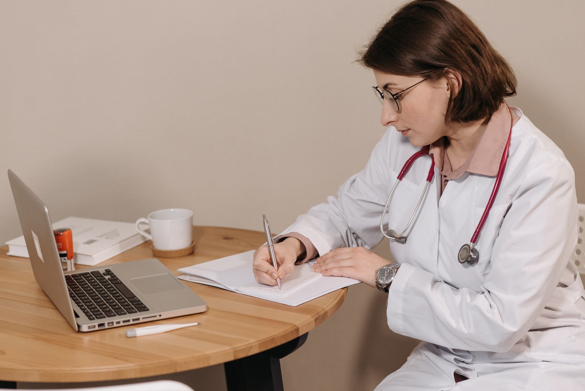 A doctor in a white coat writing notes while consulting a laptop.