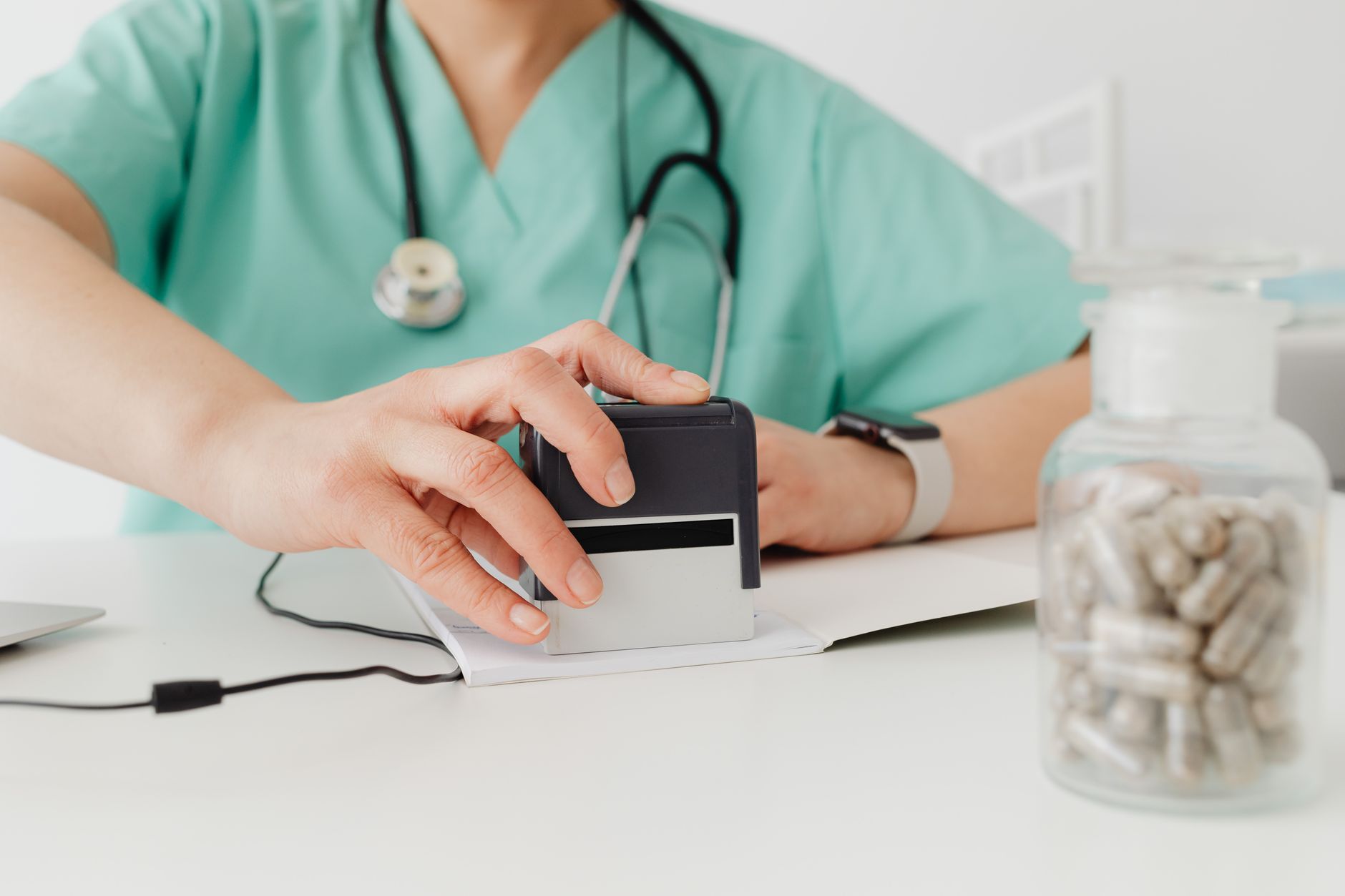 Close-up of a nurse in scrubs using a device while pills are visible on the desk.