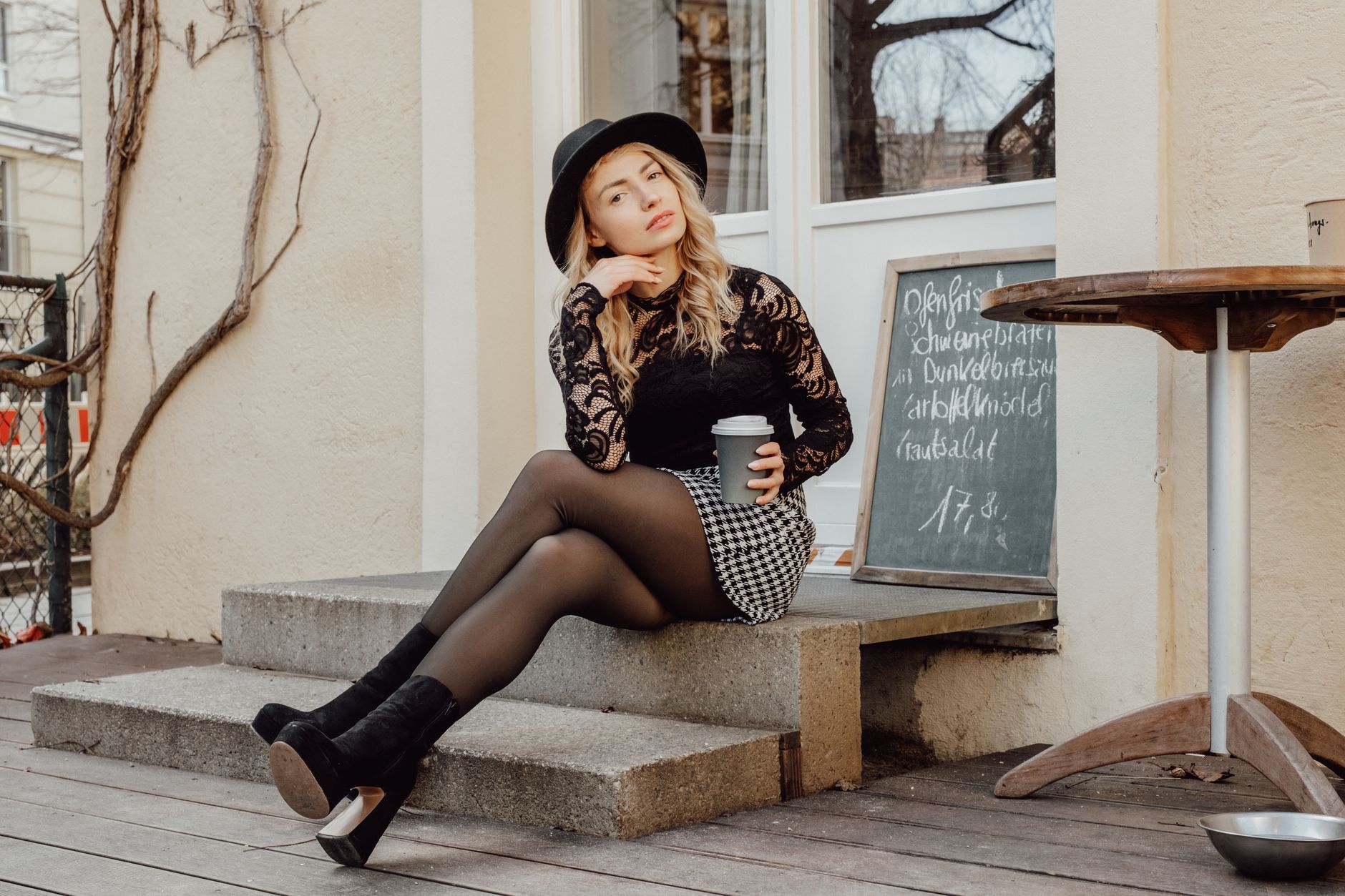 A stylish woman in a black lace top and checkered skirt sits on steps, holding coffee.